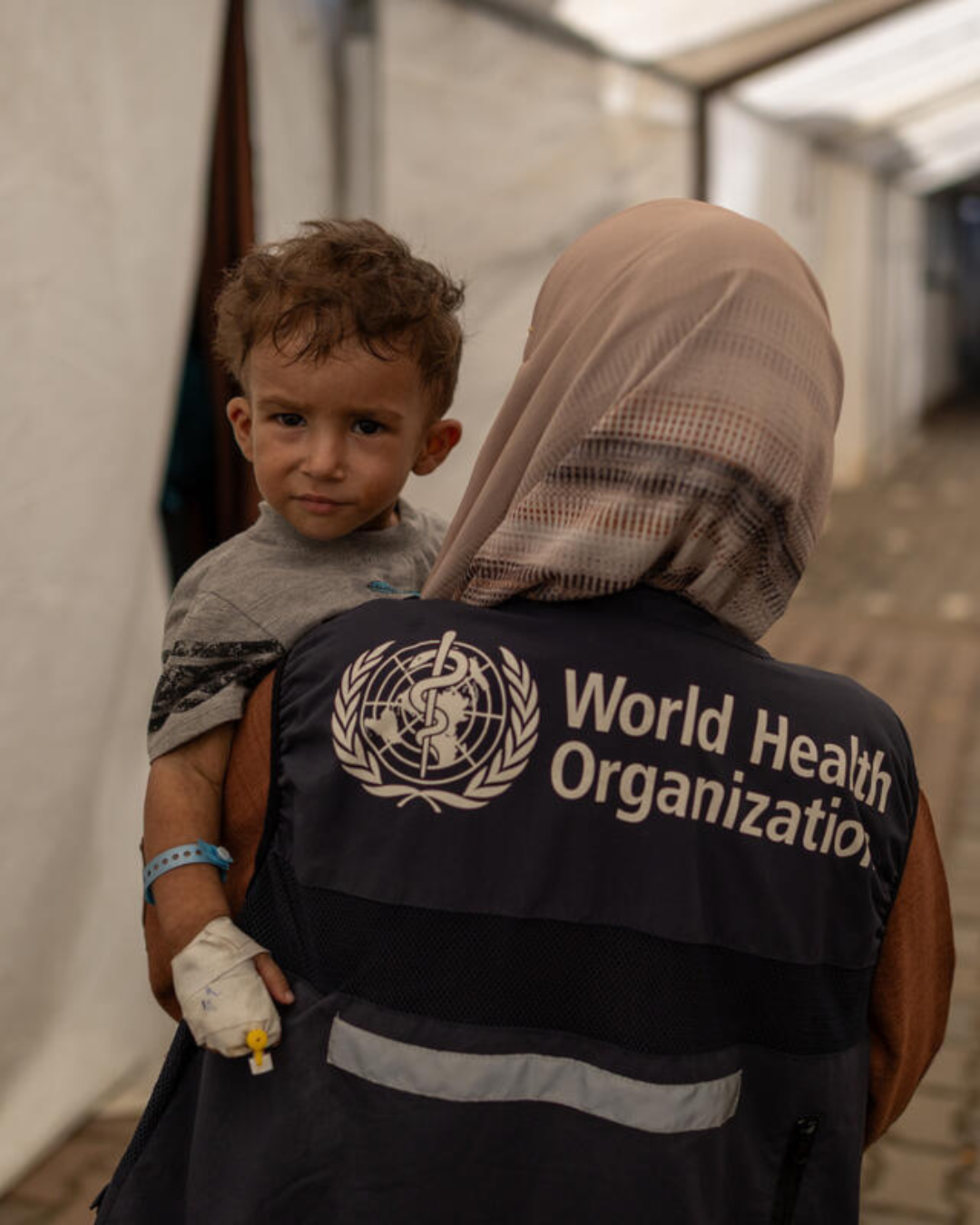 Child with a bandaged hand being carried by a woman wearing a World Health Organization vest and a beige headscarf inside a medical tent.