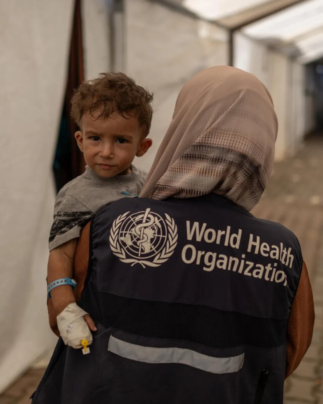 Child with a bandaged hand being carried by a woman wearing a World Health Organization vest and a beige headscarf inside a medical tent.