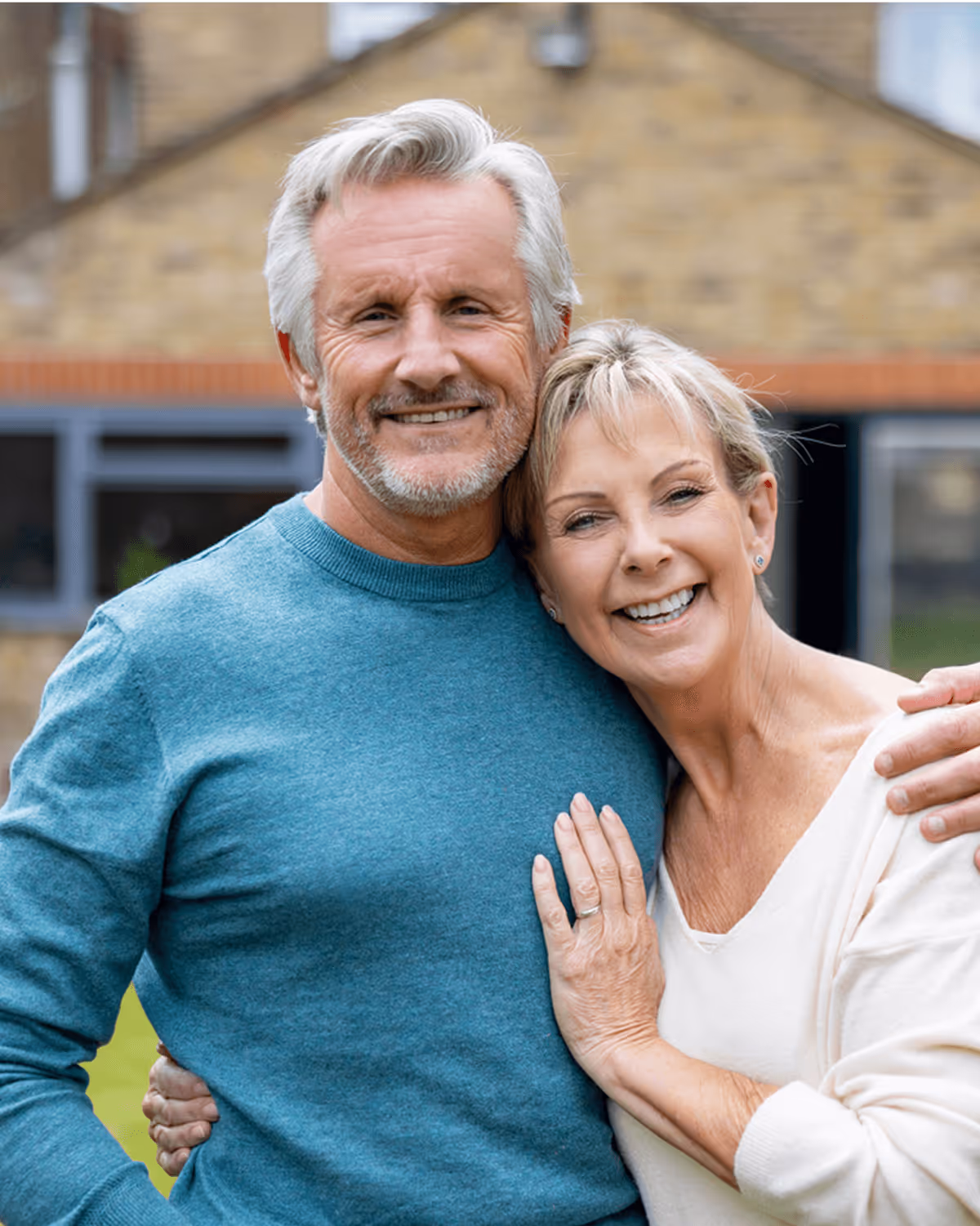 Älteres Paar umarmt sich lächelnd im Garten vor einem Haus, sonniger Tag, entspannt und glücklich.