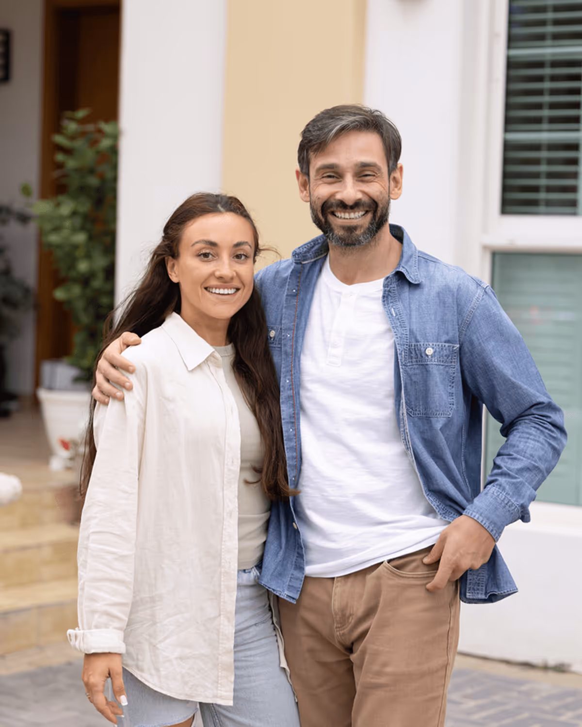 Lächelndes Paar in Freizeitkleidung steht draußen vor einem Gebäude, er trägt ein Jeanshemd, sie eine weiße Bluse.