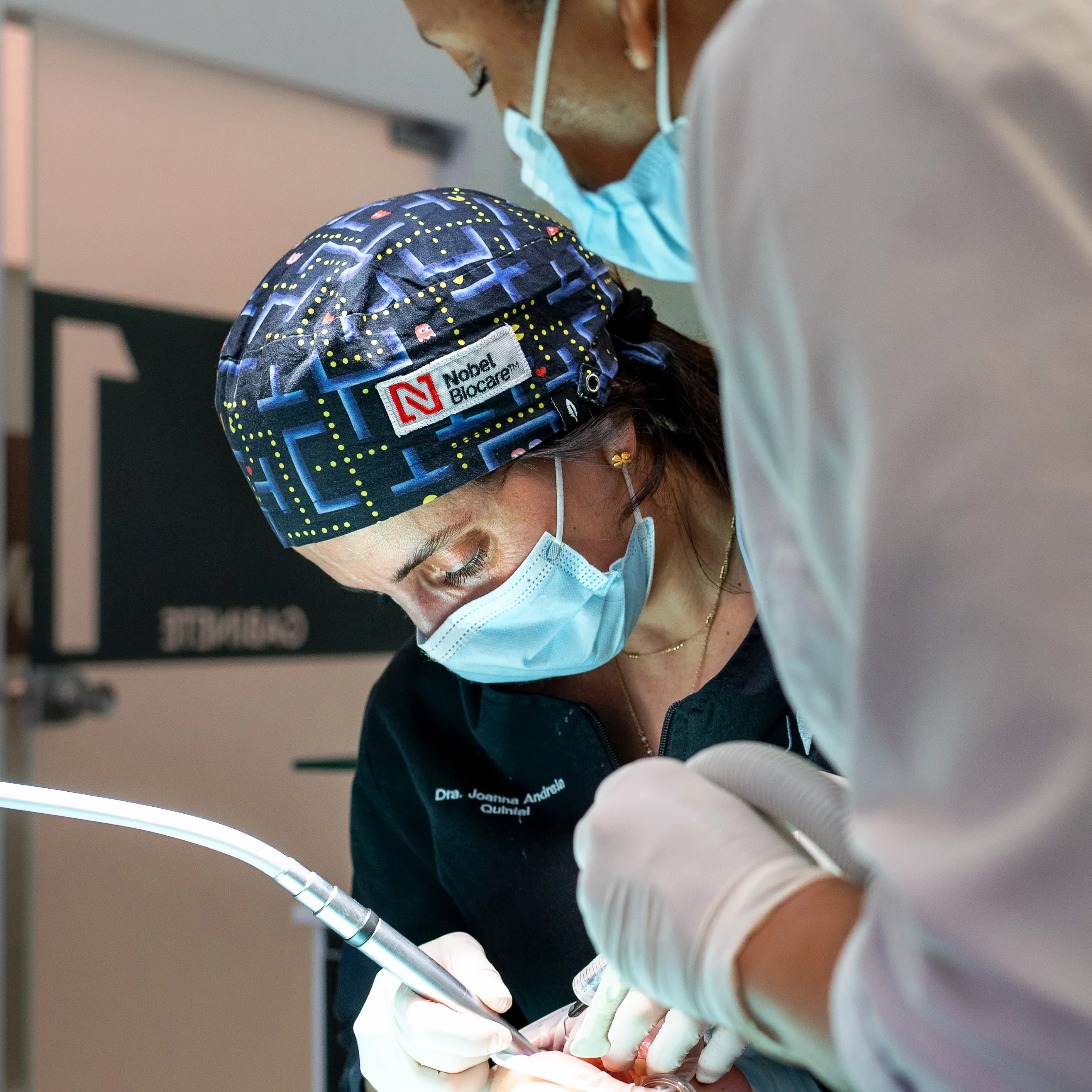 Dentista usando touca colorida e máscara cirúrgica enquanto realiza um procedimento odontológico assistido por um colega.