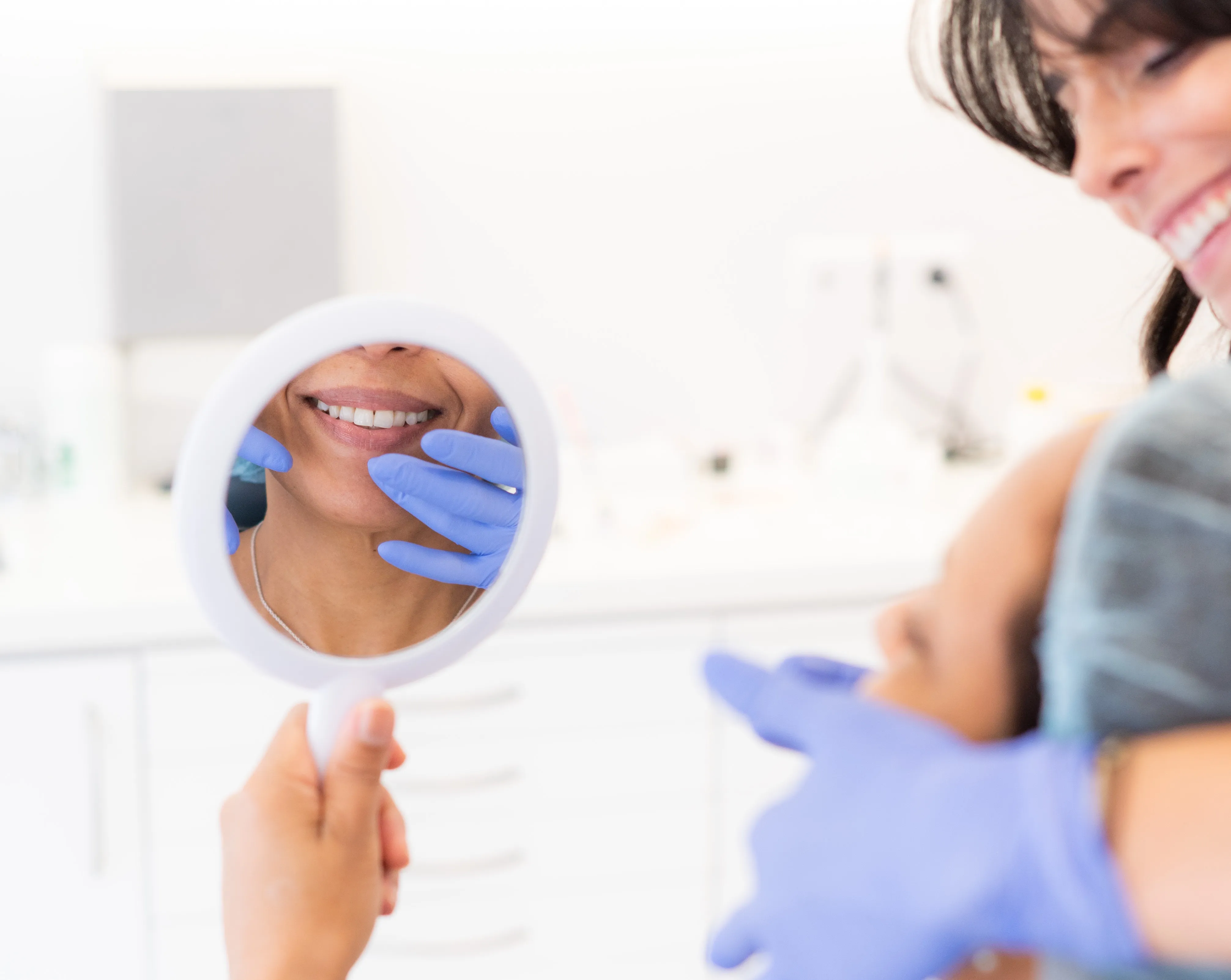 Paciente sorridente a segurar um espelho enquanto profissional com luvas azuis examina os lábios.