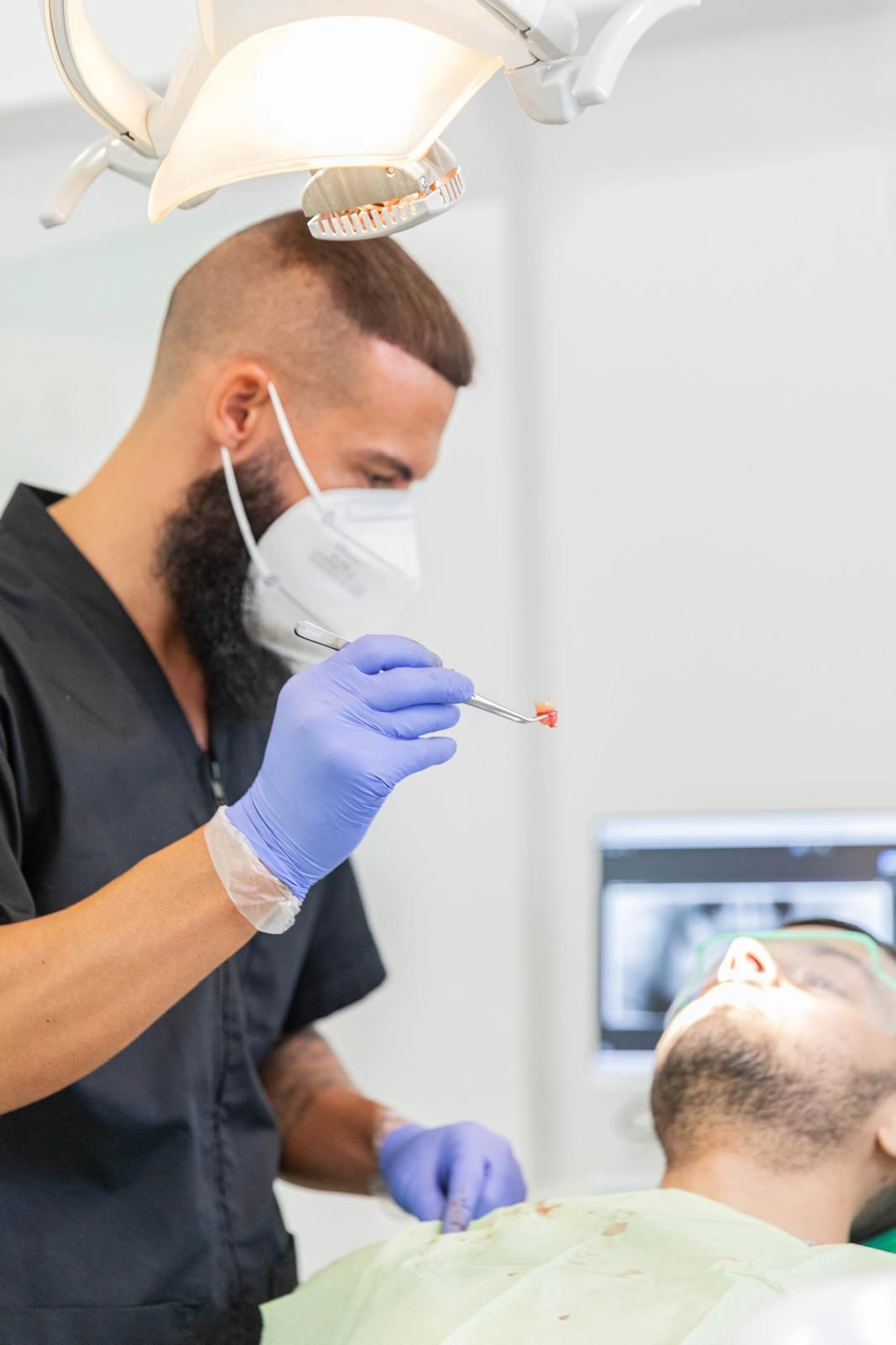 Médico Dentista com máscara e luvas segurando um dente extraído próximo a um paciente deitado na cadeira odontológica.