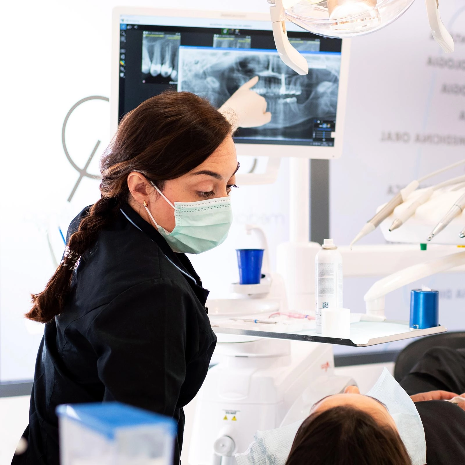 Médica dentista, a usar máscara, observa o paciente enquanto aponta para um raio-x dentário num monitor no consultório odontológico.