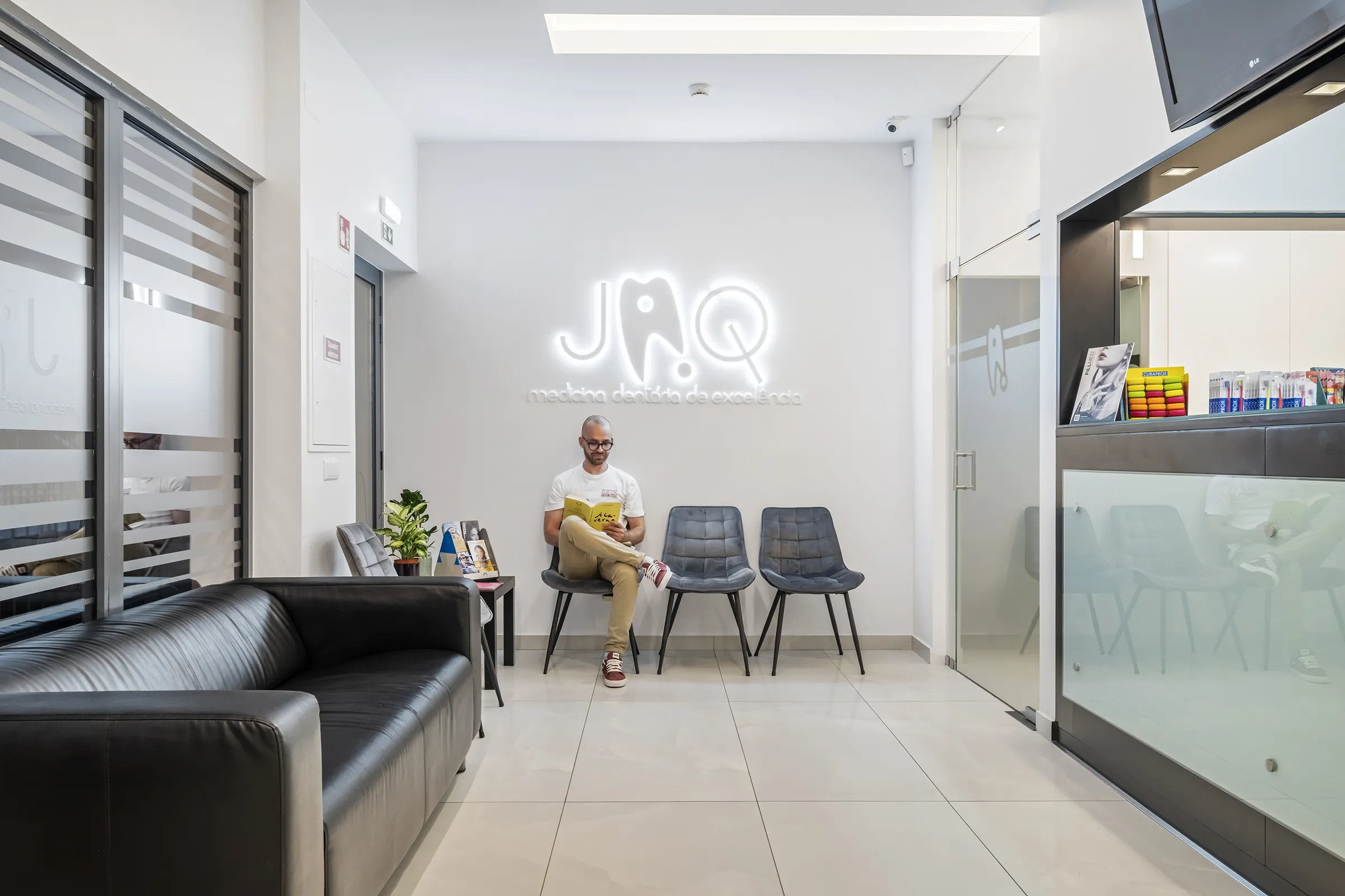 Sala de espera da JAQ Clínica Dentária, moderna e iluminada com cadeiras cinzas, sofá preto. Paciente sentado a ler uma revista. 