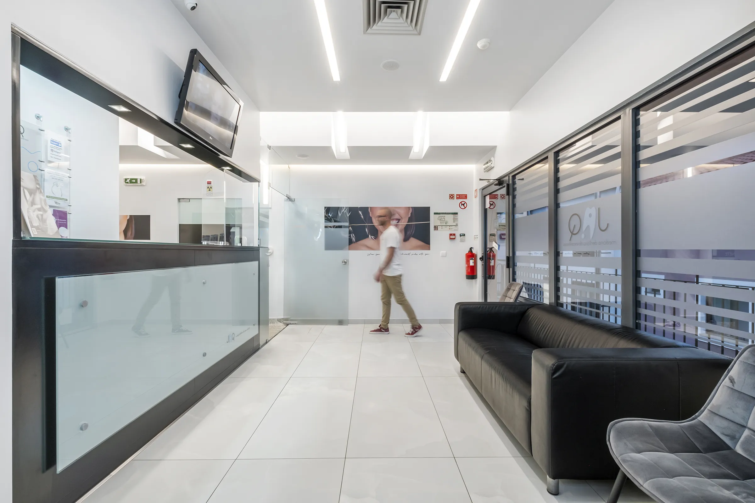 Recepção da JAQ Clínica Dentária: moderna com sofá preto, cadeiras cinzentas, balcão com vidro fosco e homem caminhando no corredor.