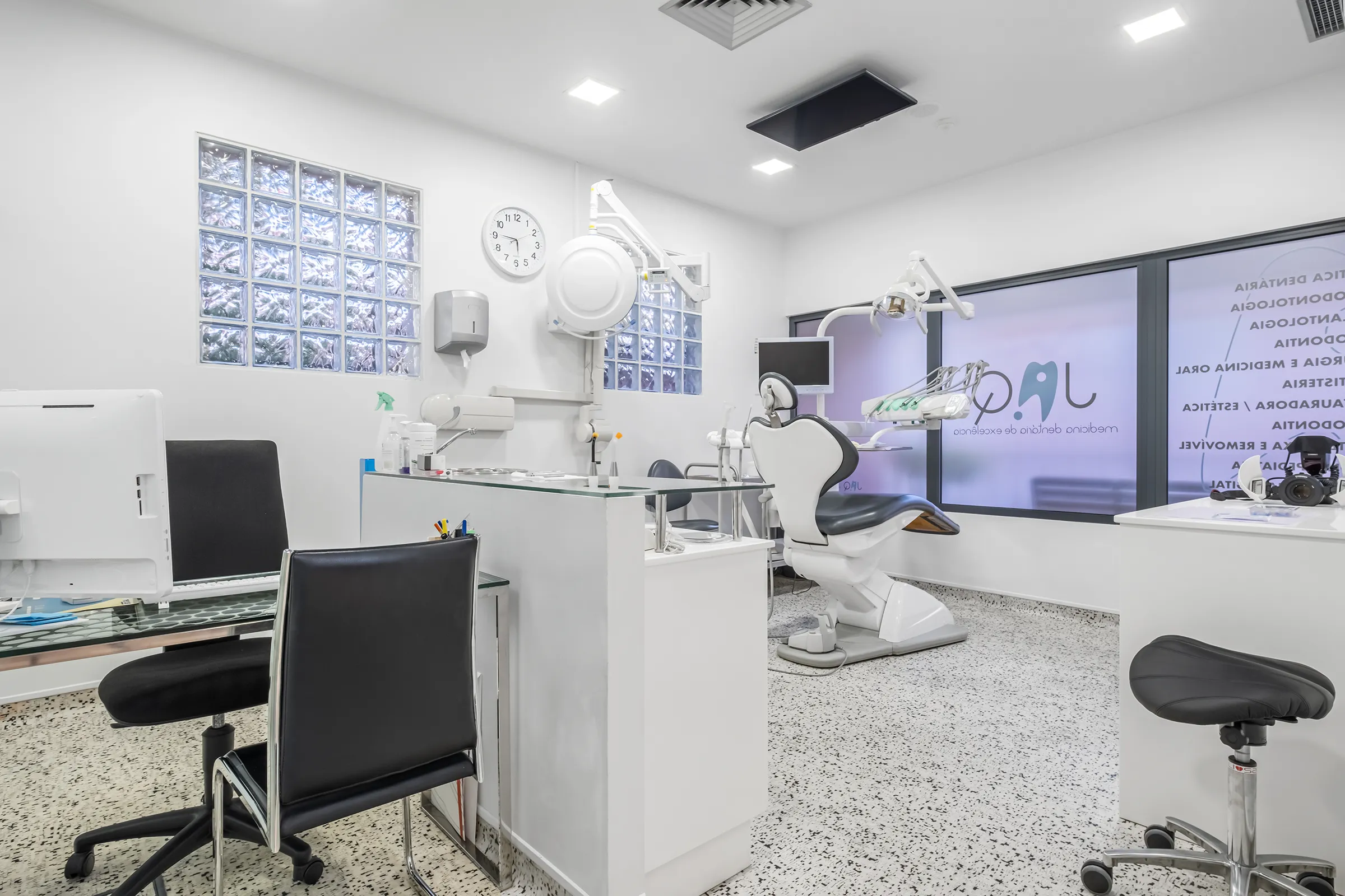 Consultório da JAQ Clínica Dentária:  amplo, moderno com cadeira de paciente, computador, cadeiras pretas e janela de vidro fosco.