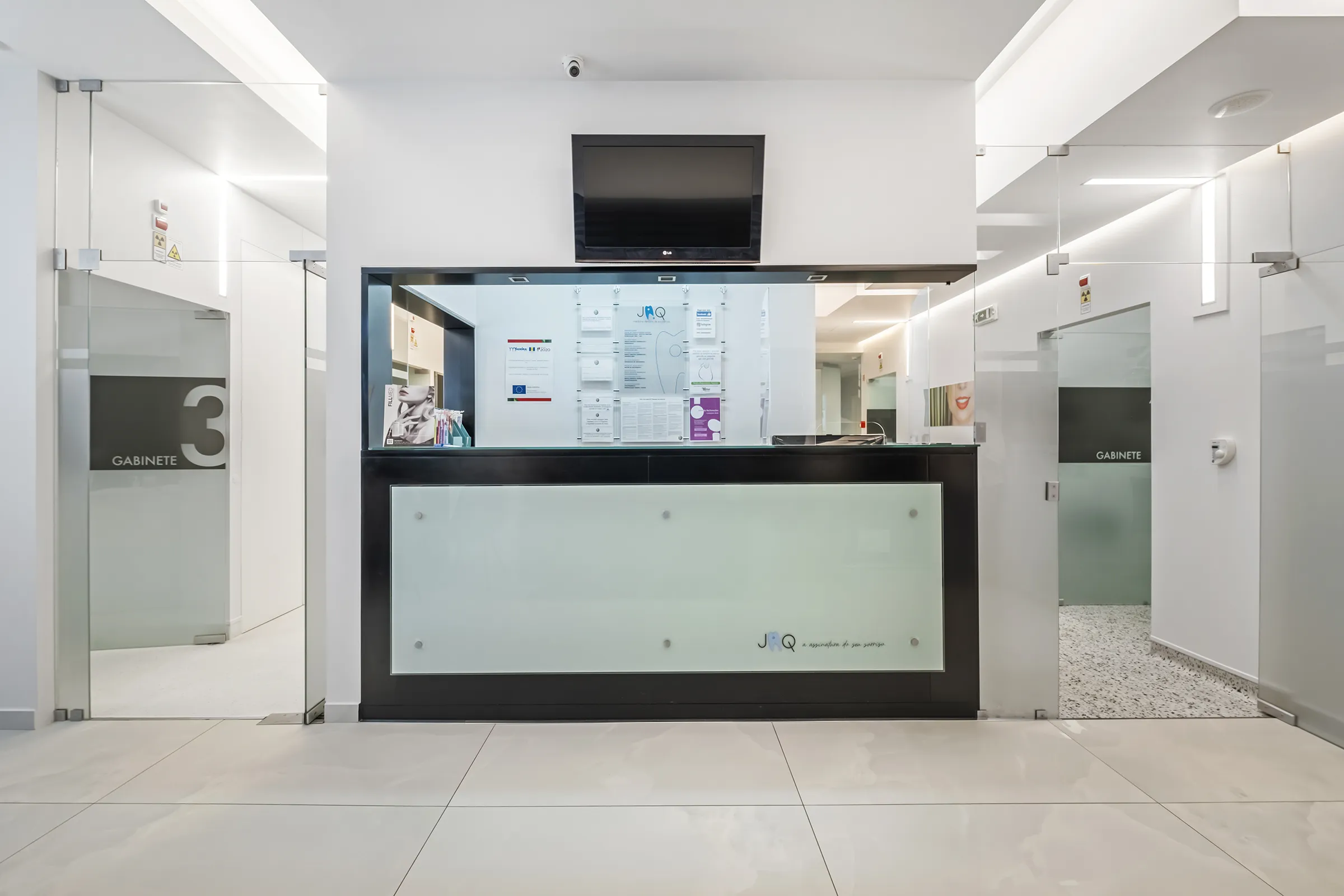 Recepção moderna da JAQ Clínica Dentária, com balcão preto e branco, monitor na parede e portas de vidro para os gabinetes.