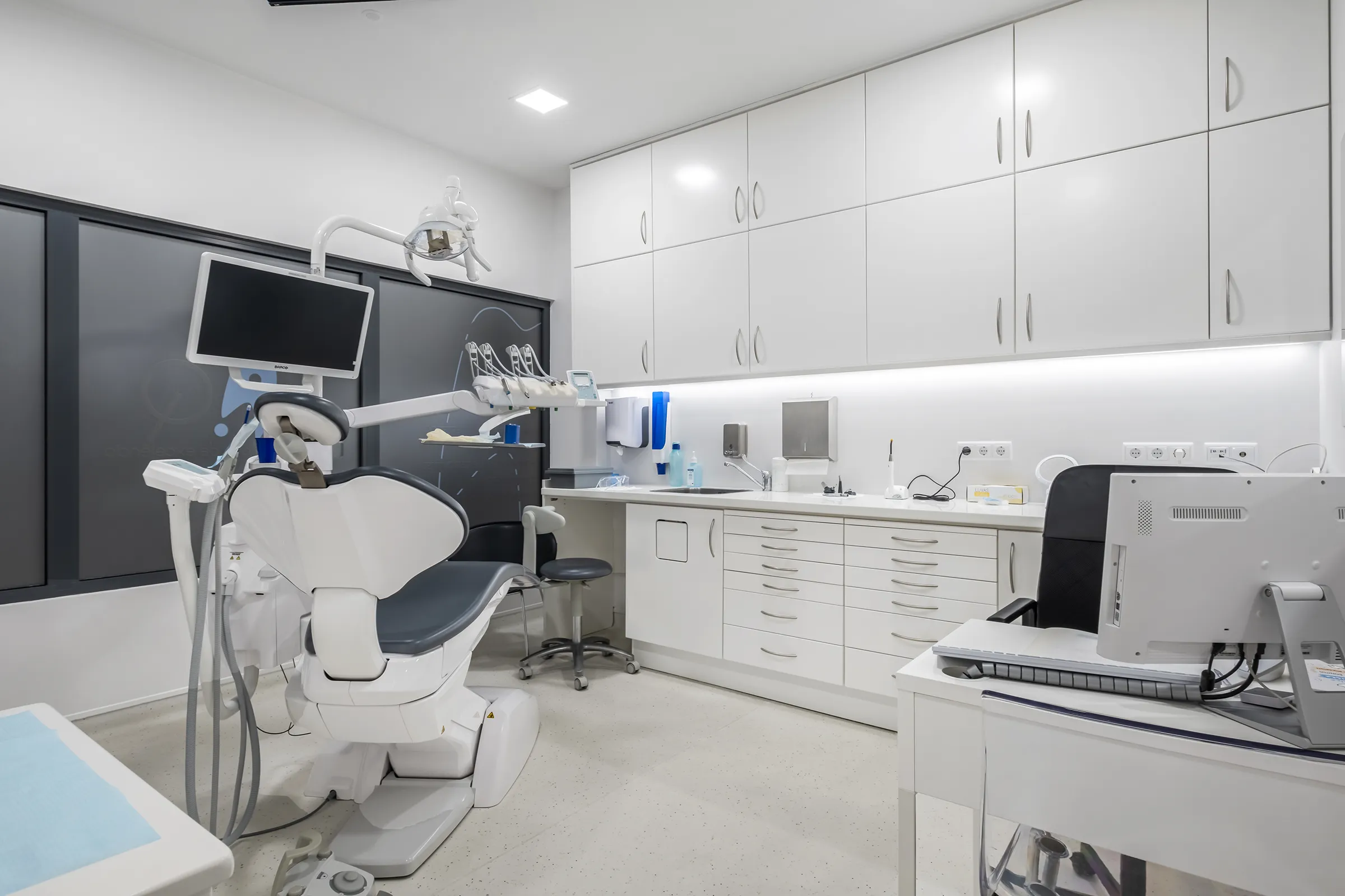 Sala da JAQ Clínica Dentária. Sala moderna com cadeira de dentista branca e preta, monitor, armários brancos e equipamento odontológico.