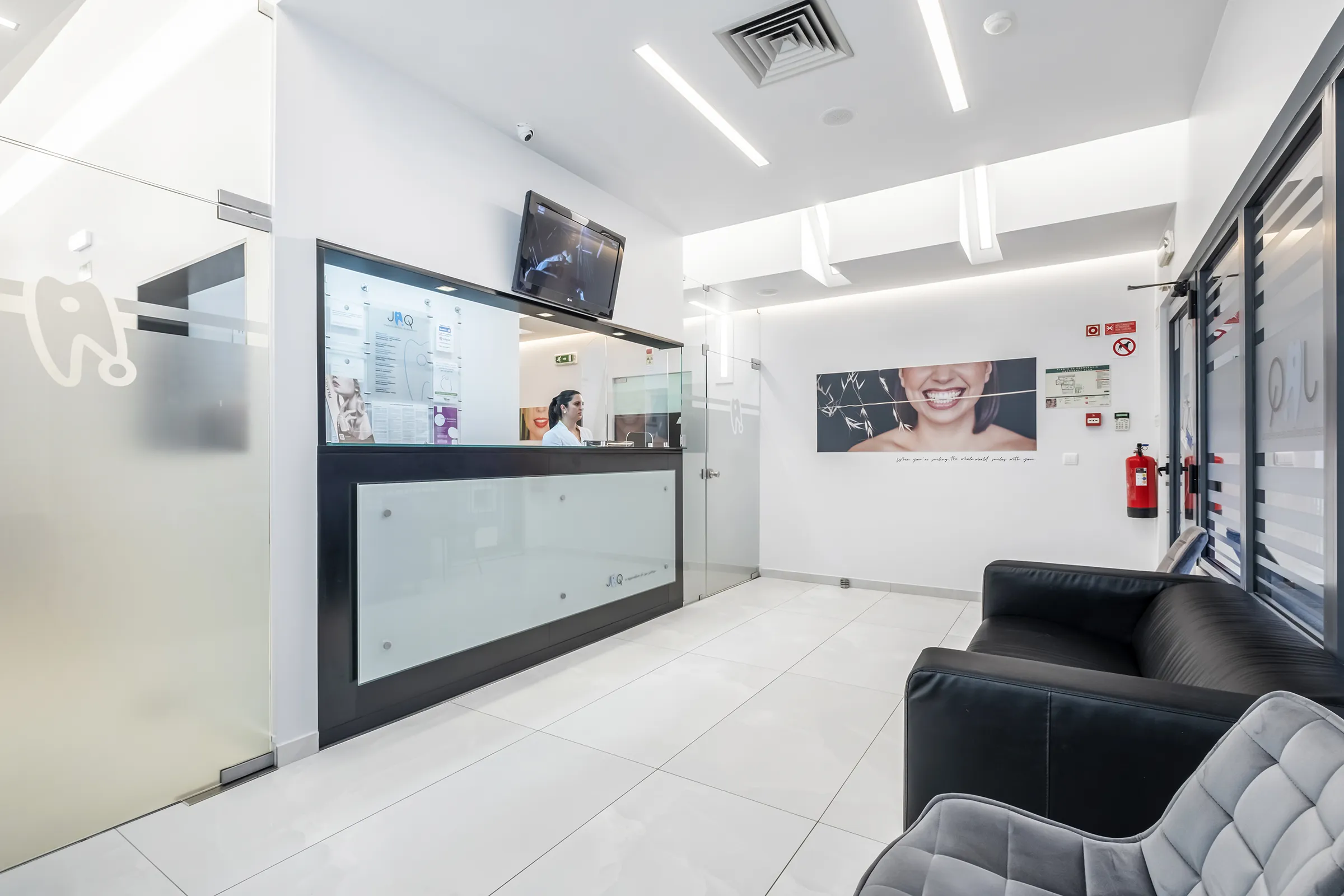 Receção da JAQ Clínica Dentária,  moderna com balcão preto e vidro fosco, TV suspensa, e sala de espera com sofá preto e cadeira cinza.