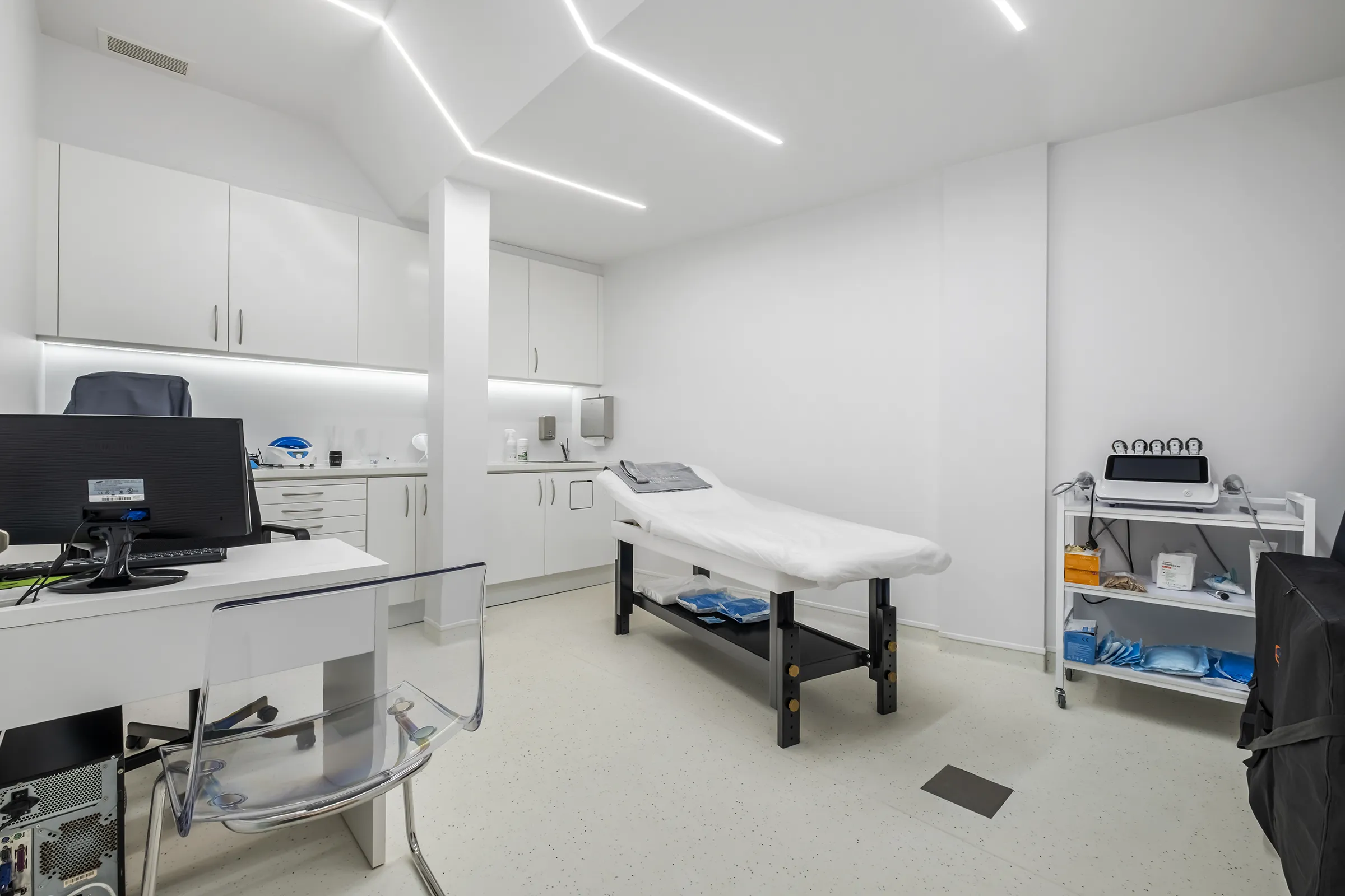 Sala clínica moderna com maca coberta, mesa com computador, cadeiras transparentes e armários brancos.