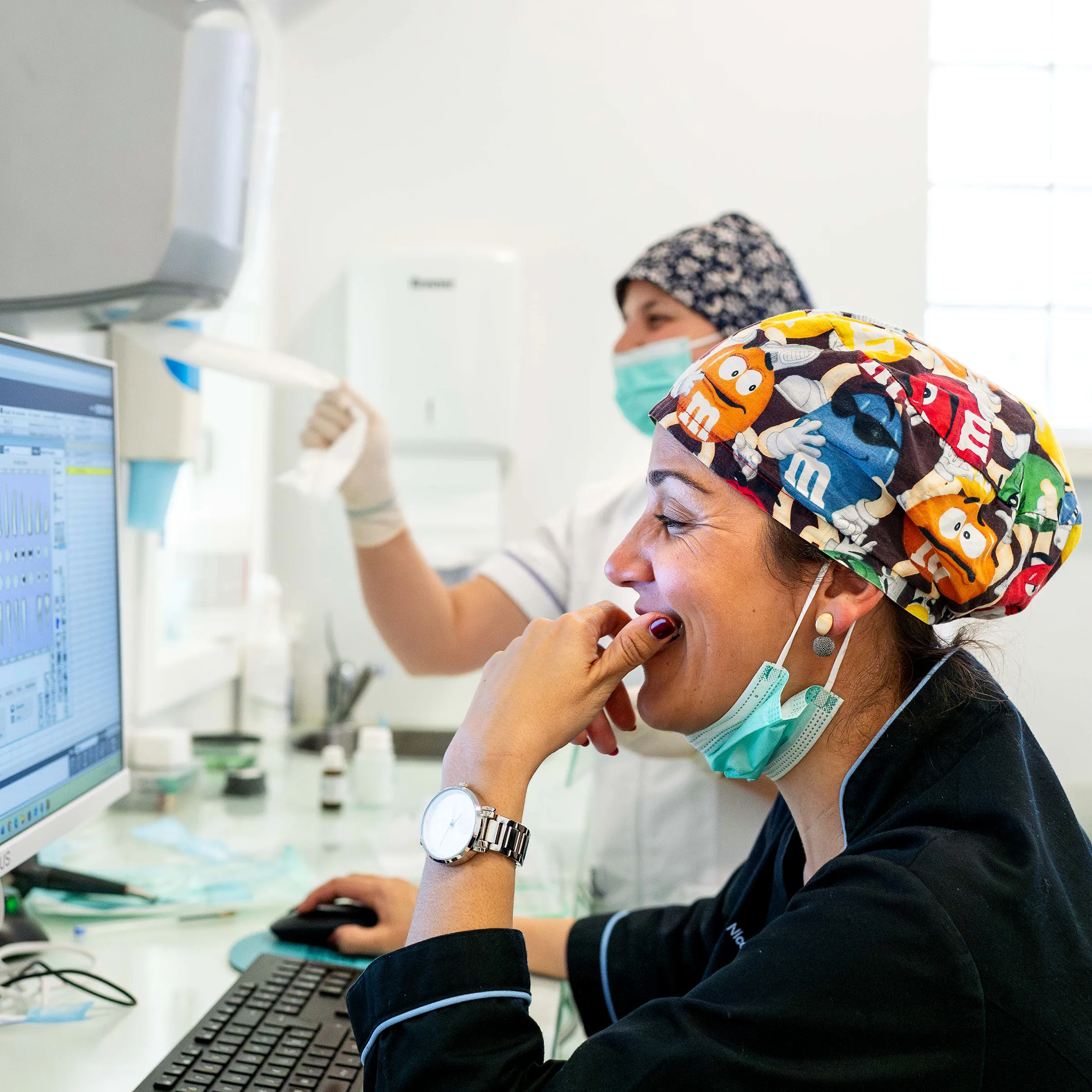 Duas profissionais de saúde sorrindo e analisando dados em computador, demonstrando bom ambiente de equipa.