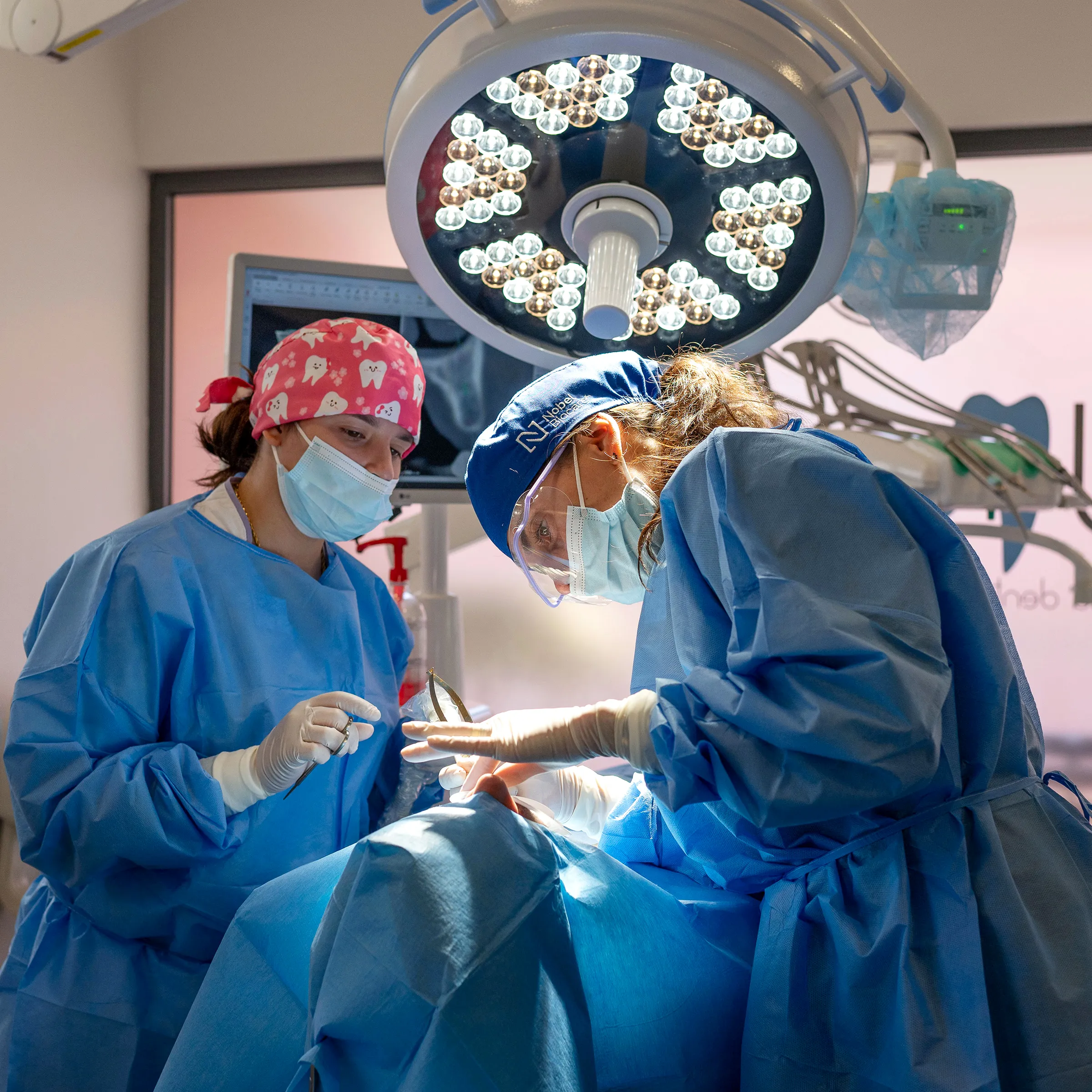 Duas dentistas usando trajes cirúrgicos e realizam um procedimento odontológico em um paciente sob luz cirúrgica.