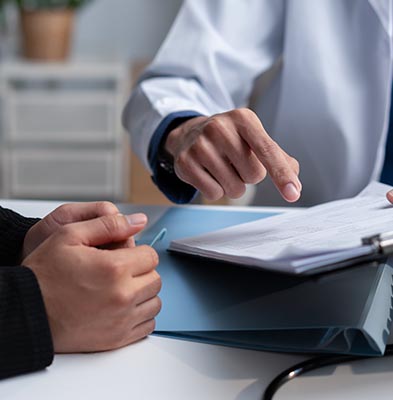 Close-up of a doctor pointing to a document on a clipboard while a patient listens, with both hands visible on a table.