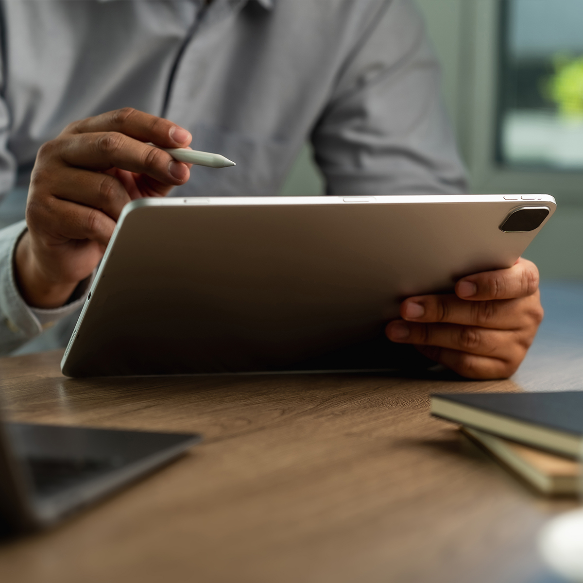 Person holding a tablet in one hand and a stylus in the other, with a laptop and notebooks on a wooden table.