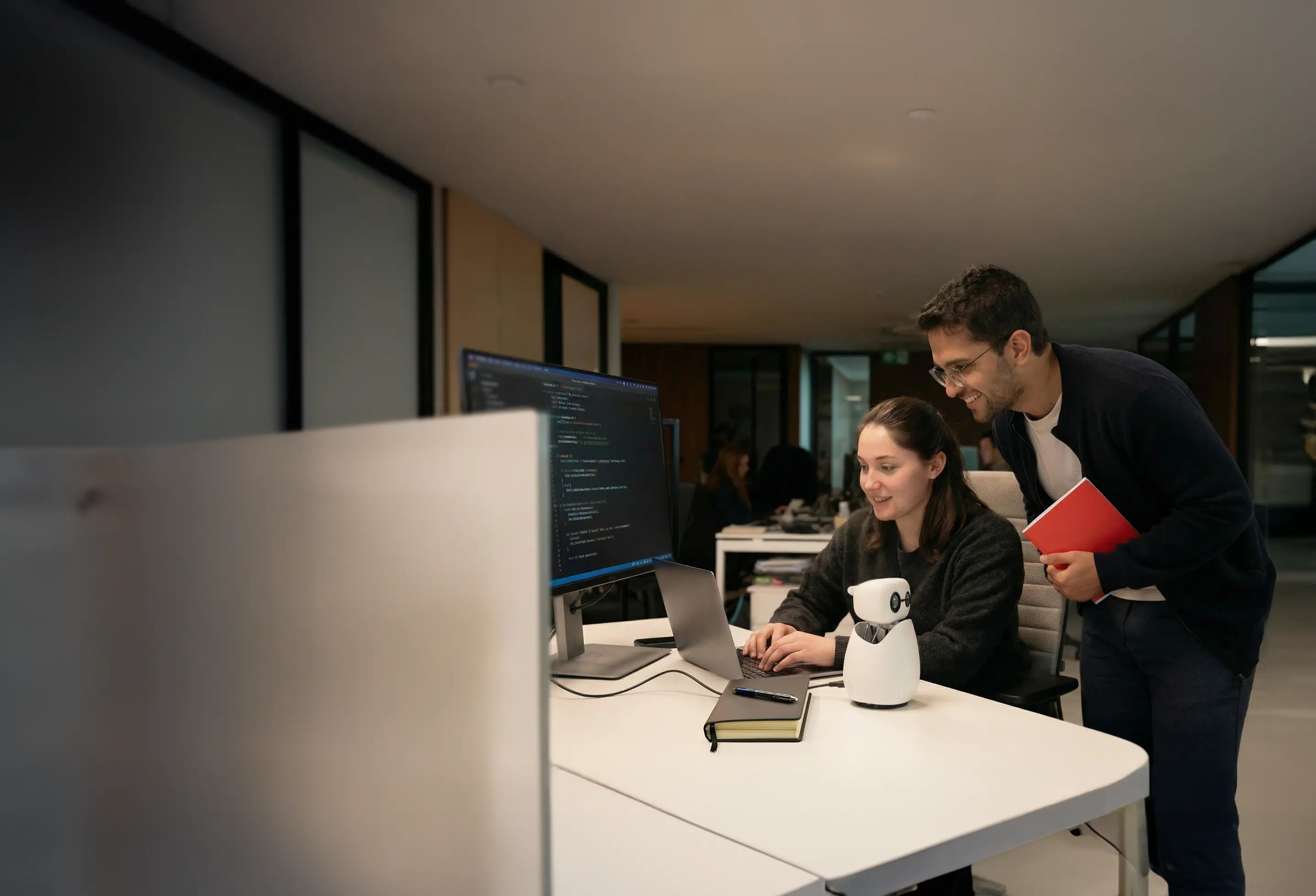 Two young professionals collaborate at a desk with a laptop, external monitor displaying code, and a small white robot.