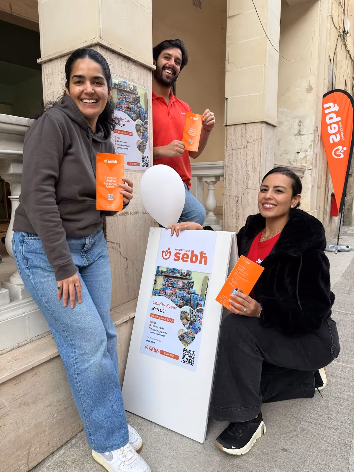 3 smiling volunteers hold flyers and a balloon while promoting a Deriv and Fondazzjoni Sebħ event outside a stone building.