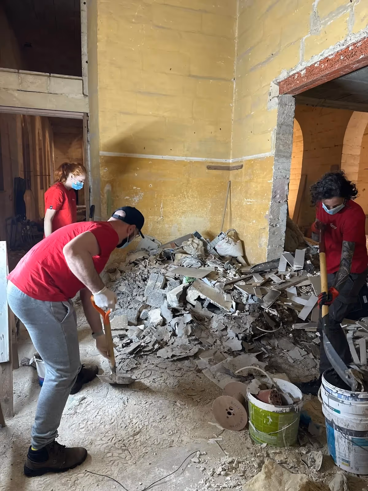 Deriv volunteers clear rubble and debris inside a damaged building as part of a renovation effort, wearing red shirts and face masks.