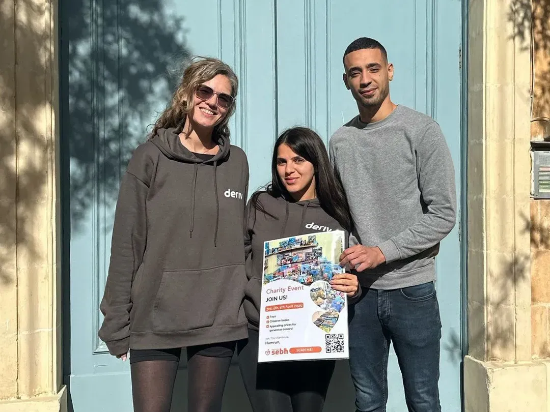 Three Deriv team members in Malta hold flyers and a poster promoting a local NGO partnership for women’s empowerment support.