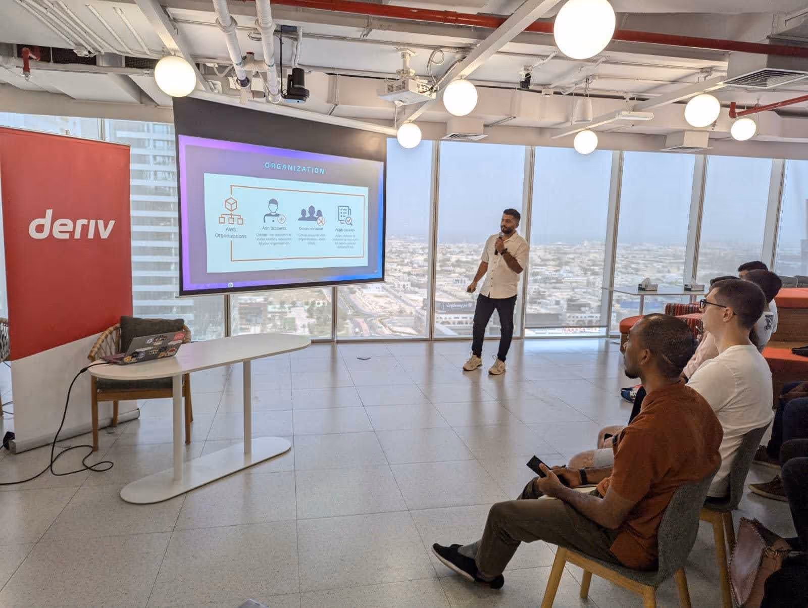 Presenter speaks about AWS Organizations at a networking event, with an audience and Deriv banner visible.