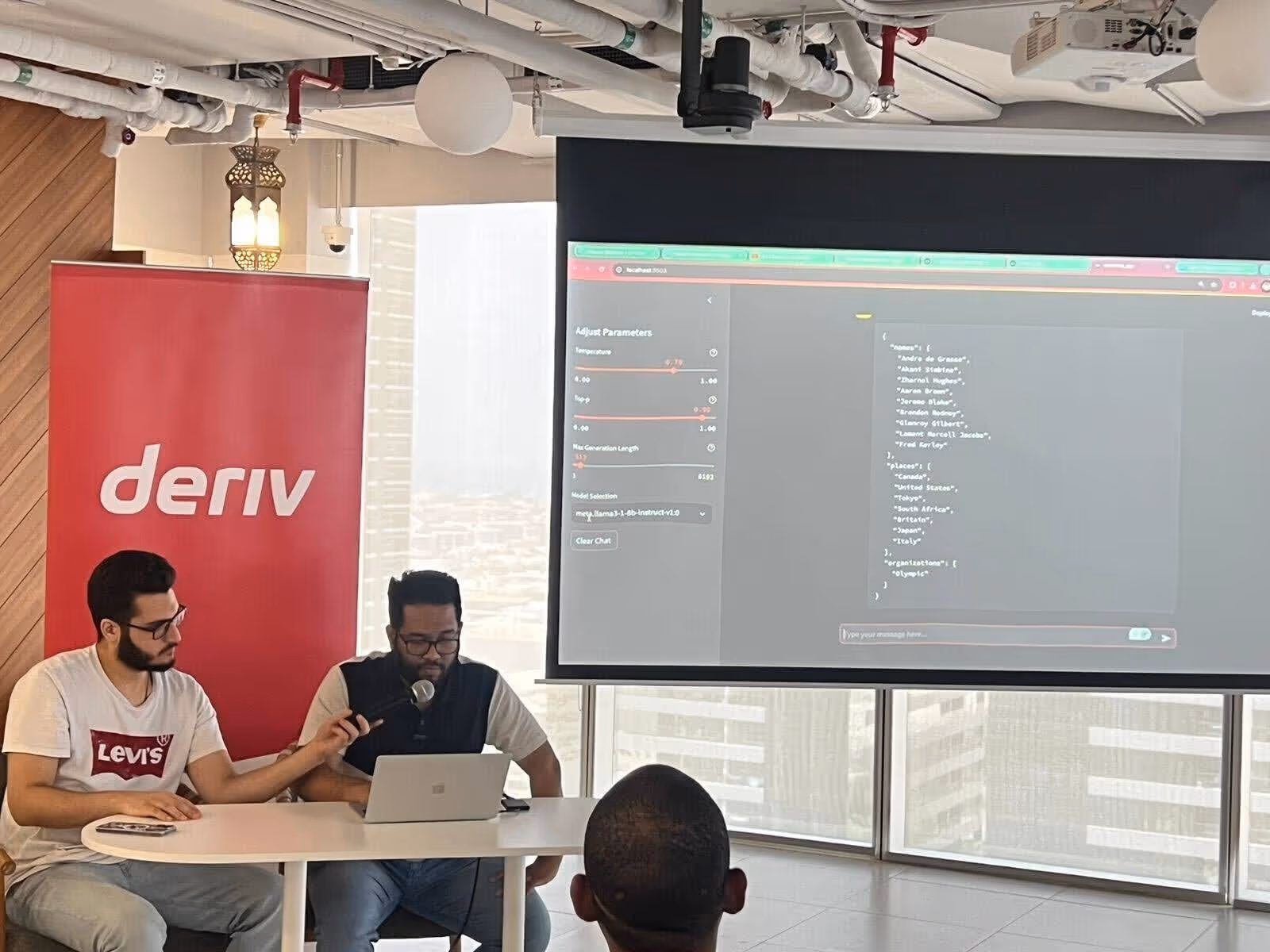 Deriv host holds a microphone while the AWS speaker presents on a laptop during an AWS community event.