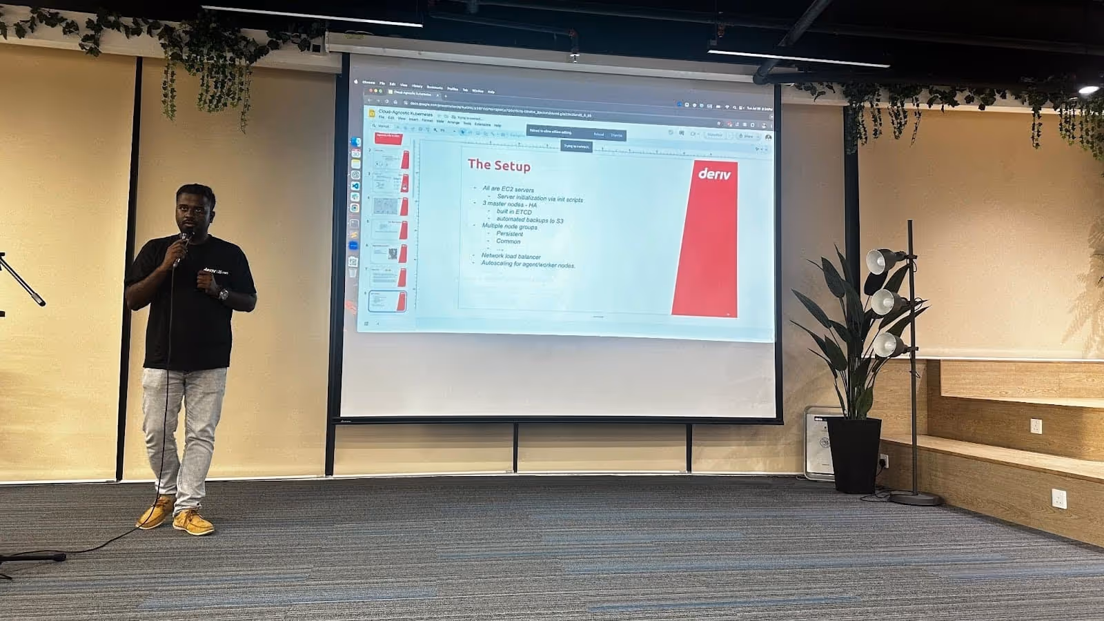 Jothimani Radhakrishnan, Deriv's Lead DevOps Engineer, speaking on stage with a presentation slide in the background.