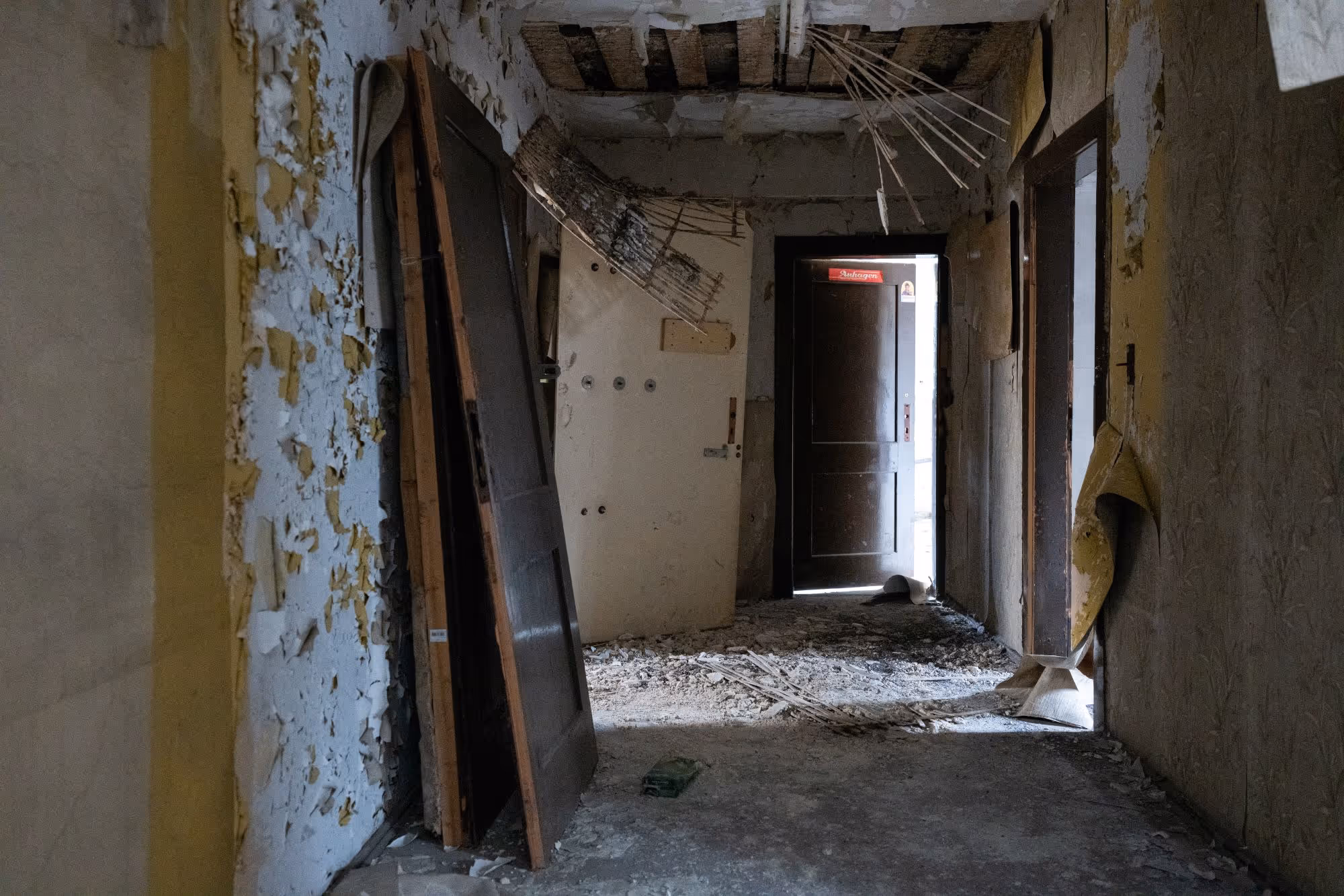 Verlassener, verfallener Flur mit abgeplatzter Wandfarbe und aufgestellten Türen, beleuchtet von Tageslicht durch eine halb geöffnete Tür am Ende.