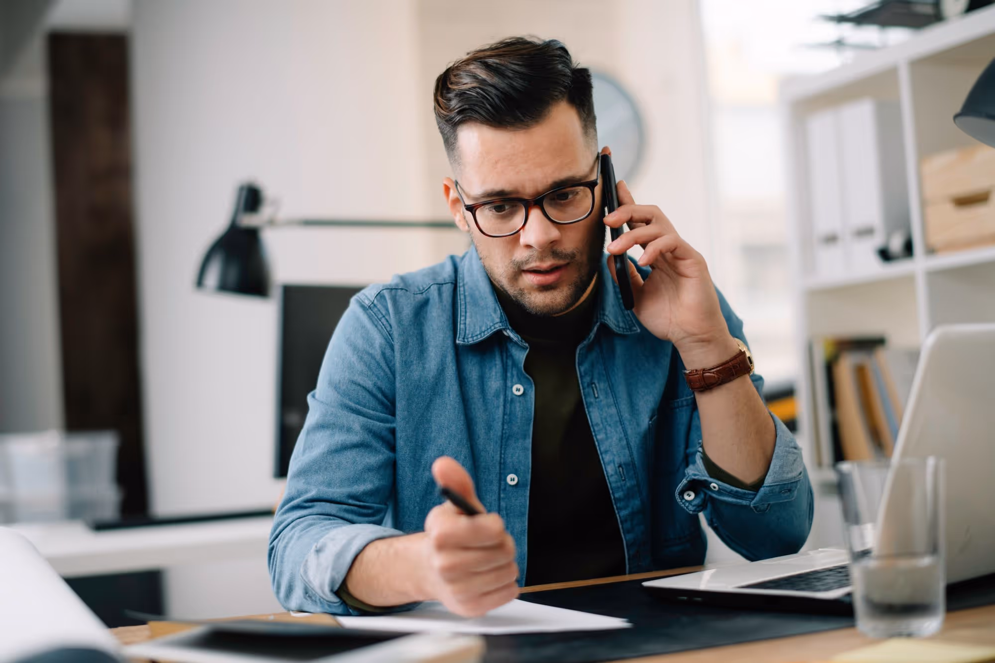Junger Mann mit Brille im Denim-Hemd telefoniert und macht sich Notizen am Schreibtisch mit Laptop.