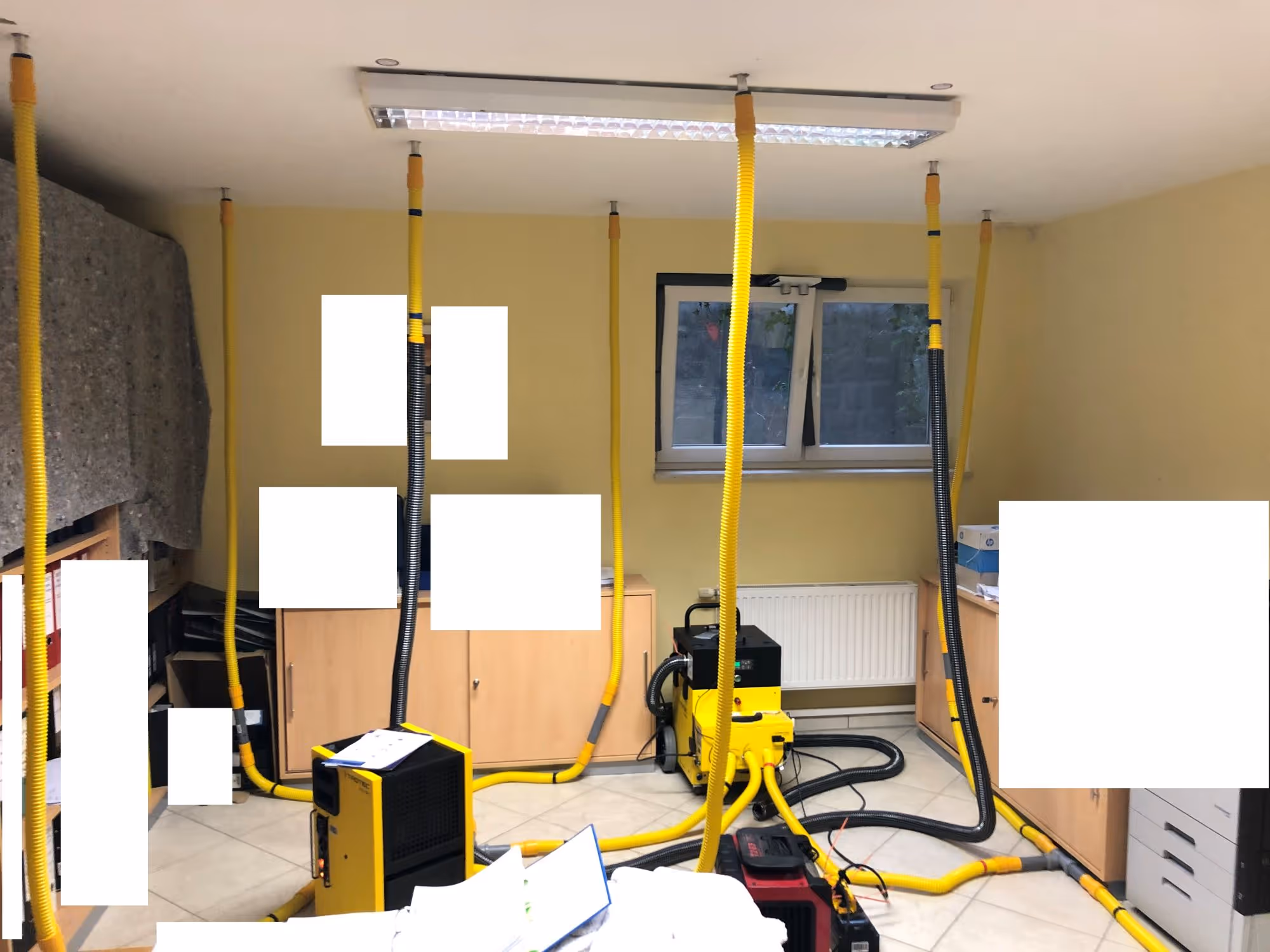 Raum mit gelben Schläuchen, die von Geräten auf dem Boden zur Decke führen, mit Aktenordnern und Schränken an den Wänden.