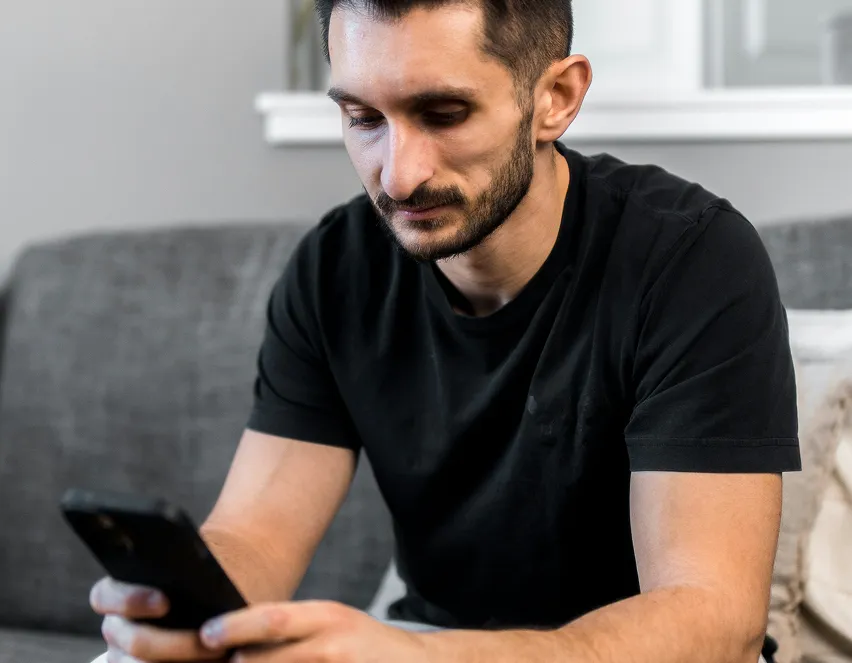Man with short dark hair and beard sitting on a gray couch, looking thoughtfully at a smartphone he holds in his hands.