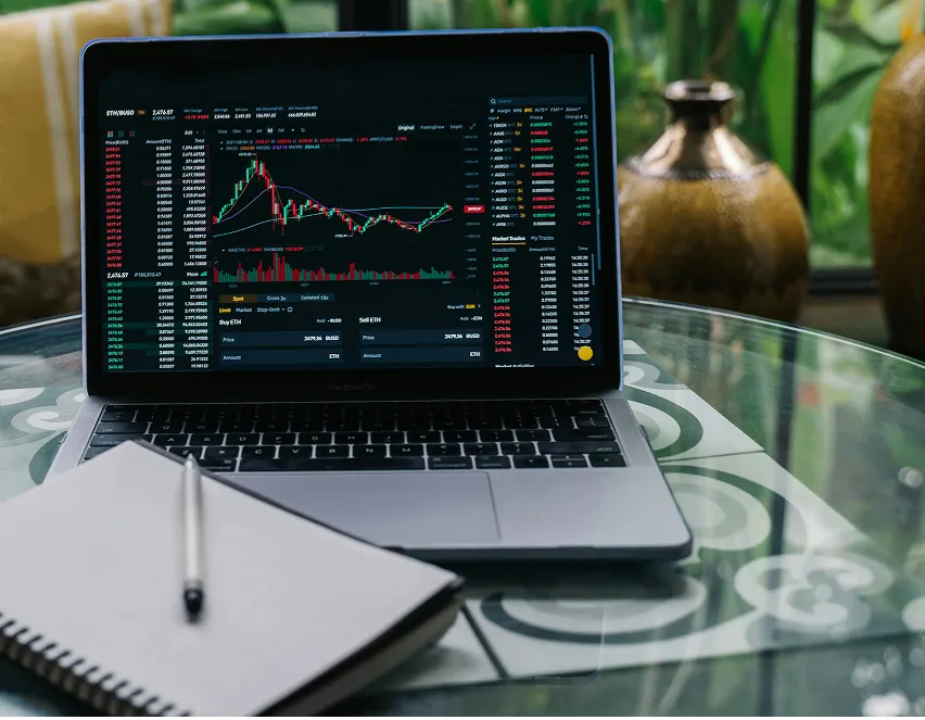 Laptop on glass table displaying a cryptocurrency trading platform with charts and market data next to a closed notebook with a pen.