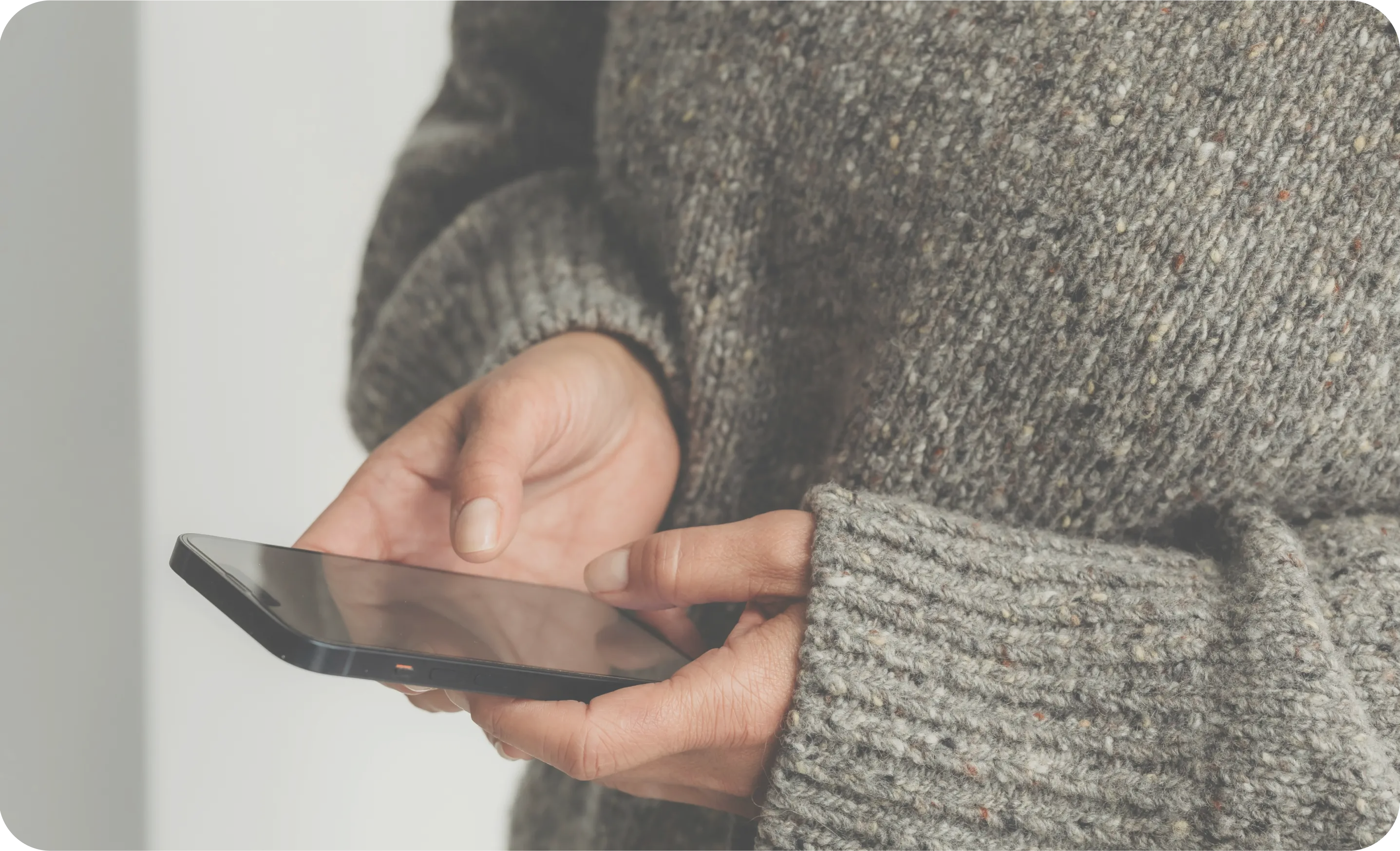 Person wearing a thick gray knitted sweater holding and using a smartphone with both hands.