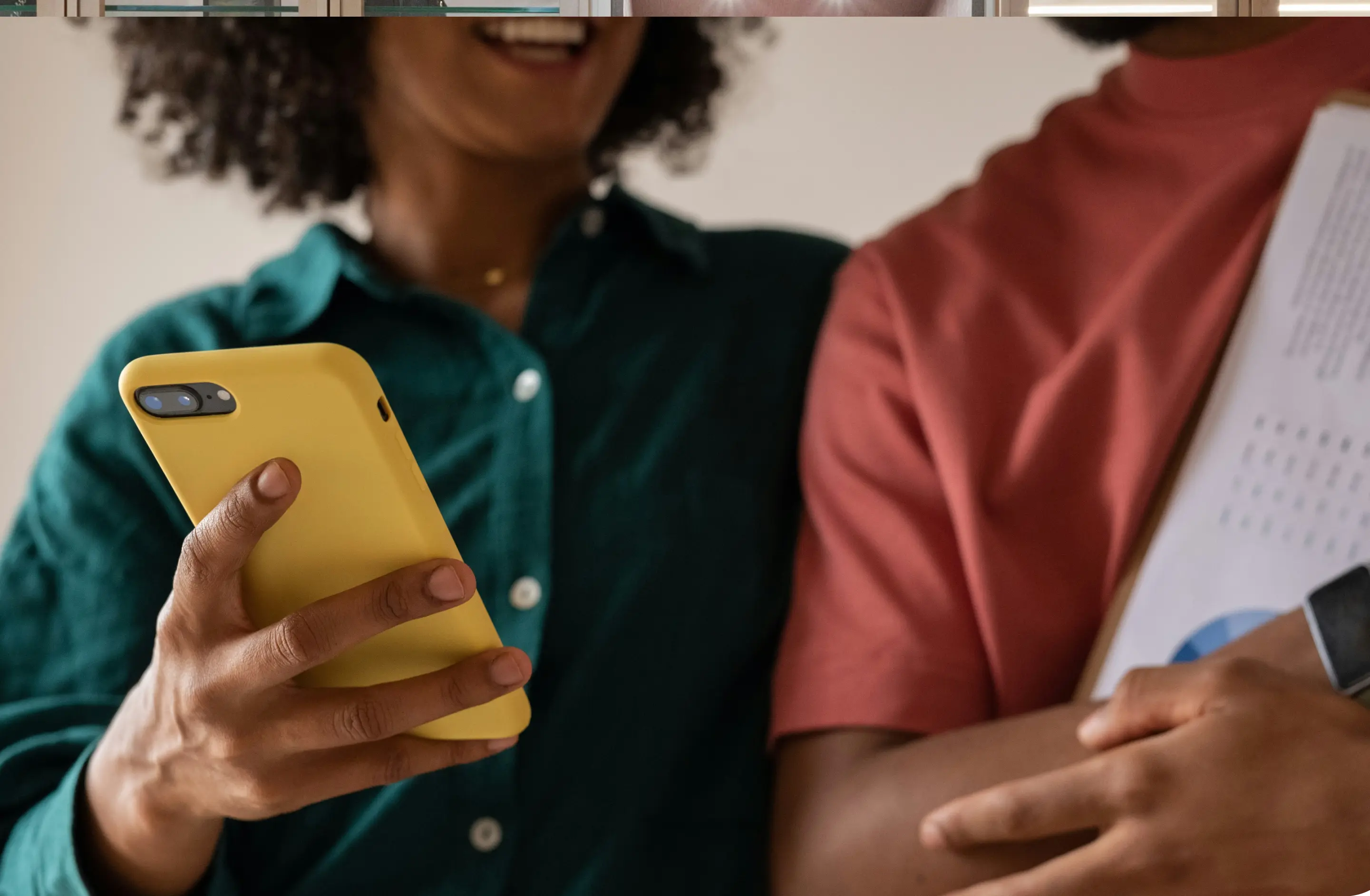 Two people smiling and looking at a yellow smartphone held by the person on the left, while the person on the right holds papers and wears a smartwatch.