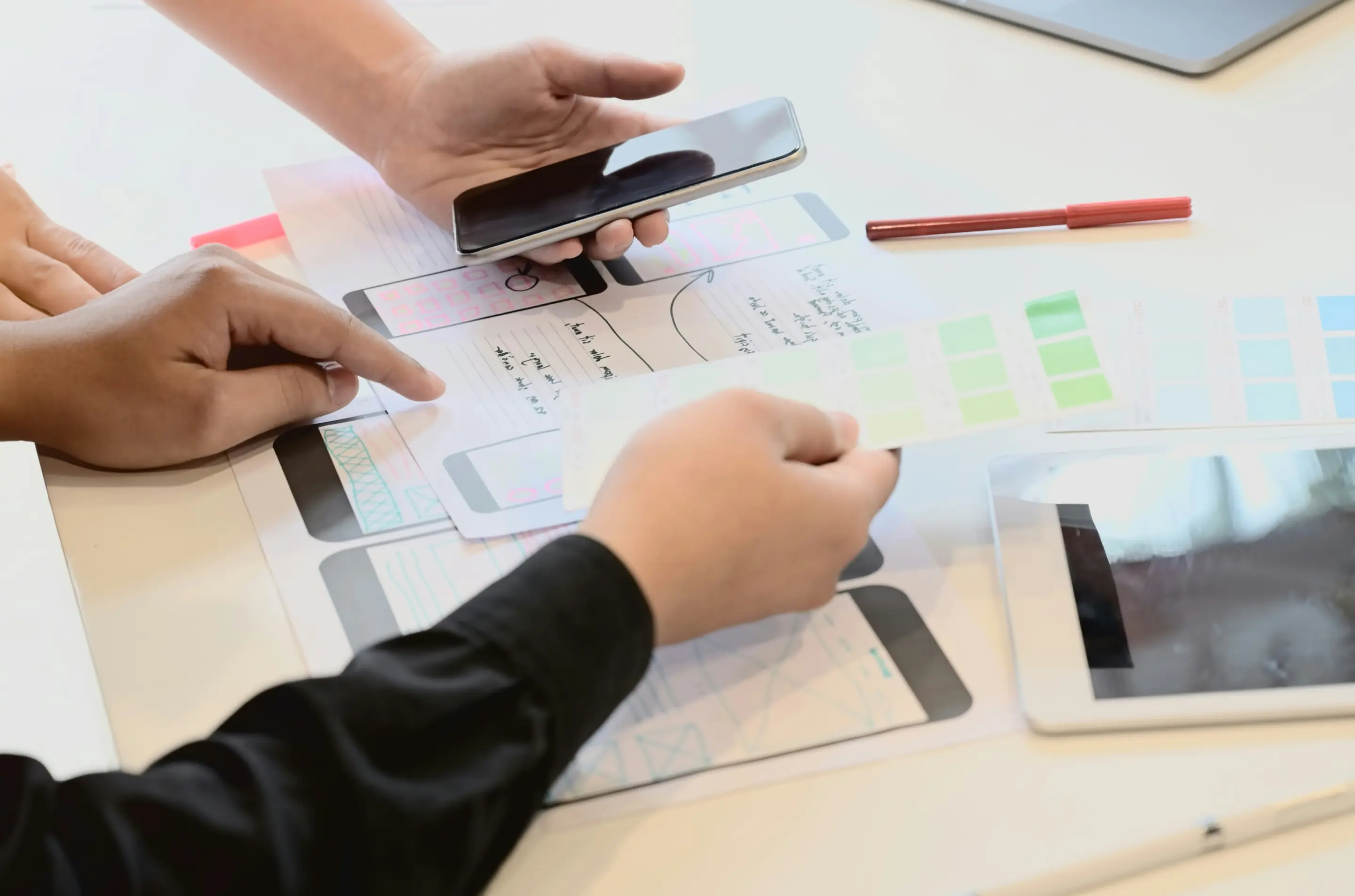 Two people reviewing smartphone app wireframes and color swatches on a table with a smartphone and tablet nearby.