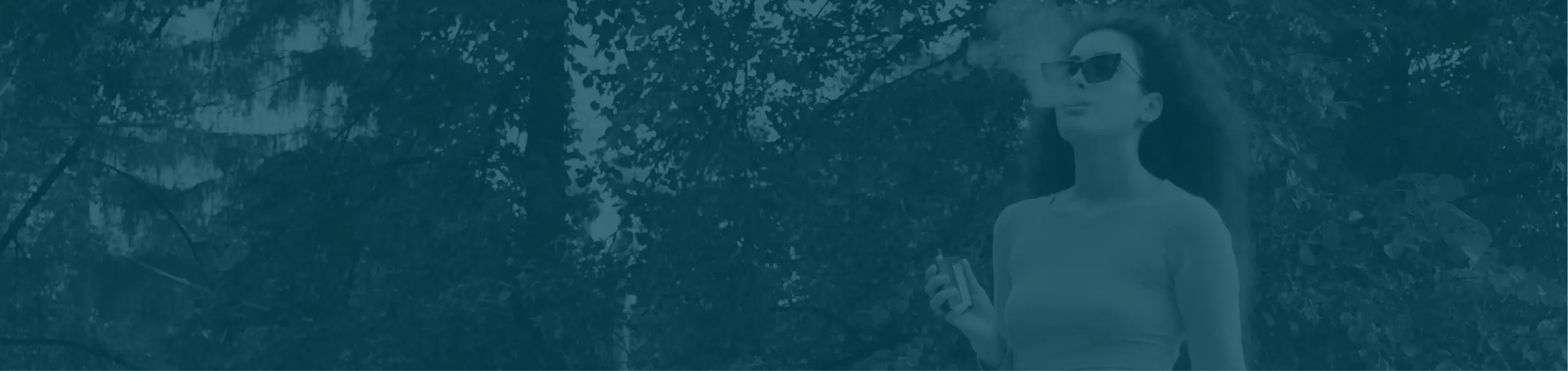 Woman wearing sunglasses exhaling vapor outside among trees while holding a vaping device.