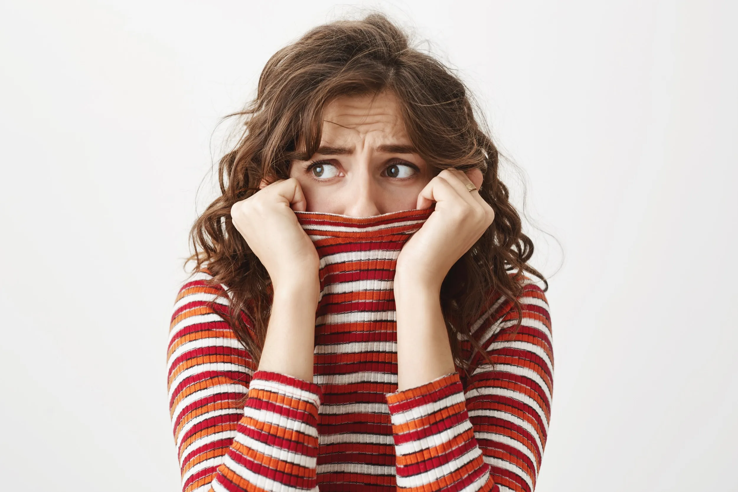 Worried woman with curly brown hair hiding her mouth behind a red, white, and orange striped sweater.