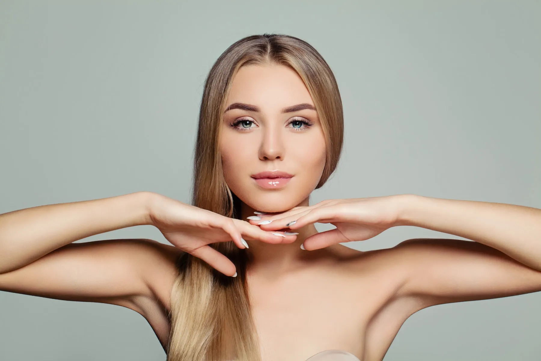 Young woman with long blonde hair posing with hands delicately framing her chin against a plain gray background.