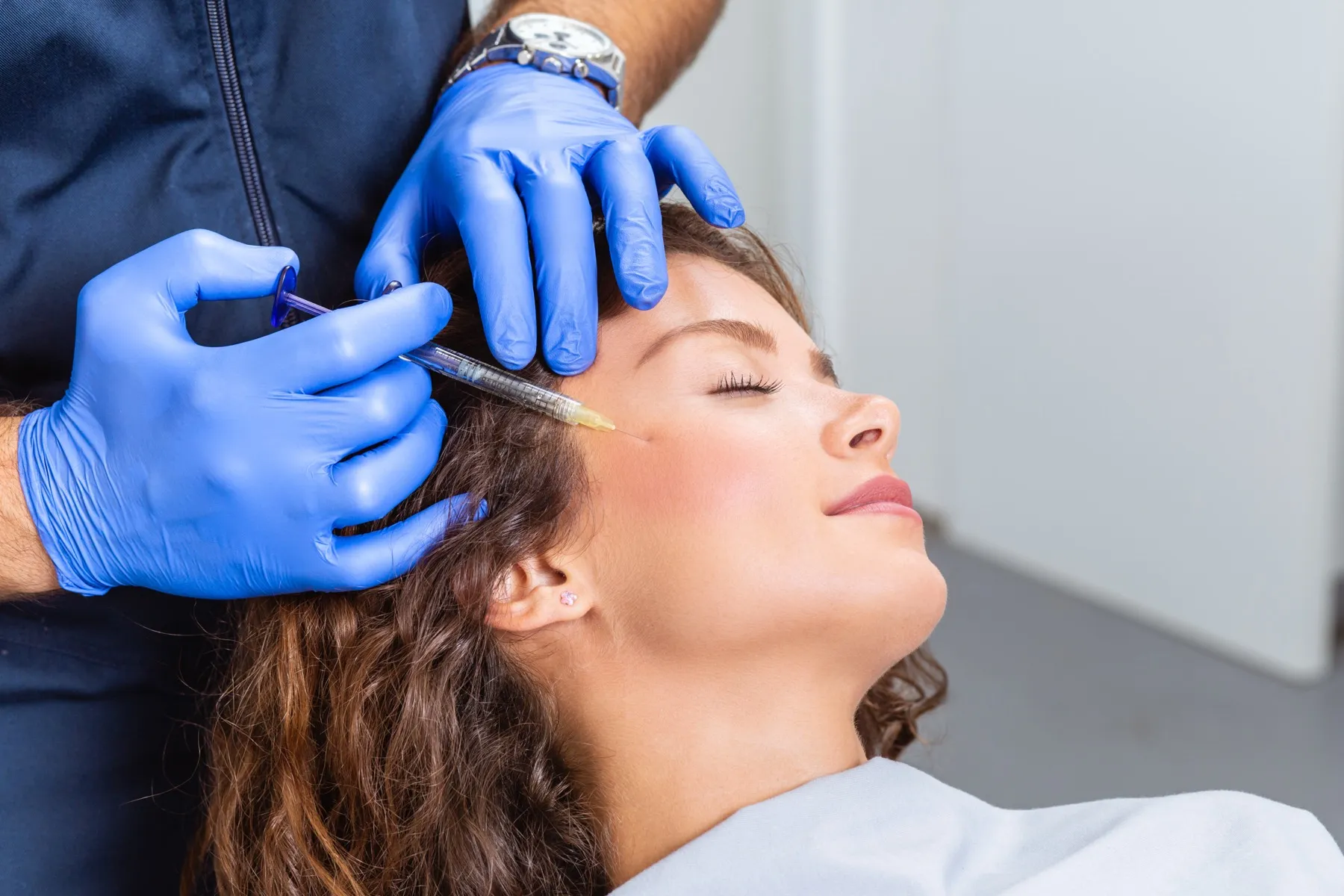 Woman receiving a cosmetic injection near her temple from a professional wearing blue gloves.