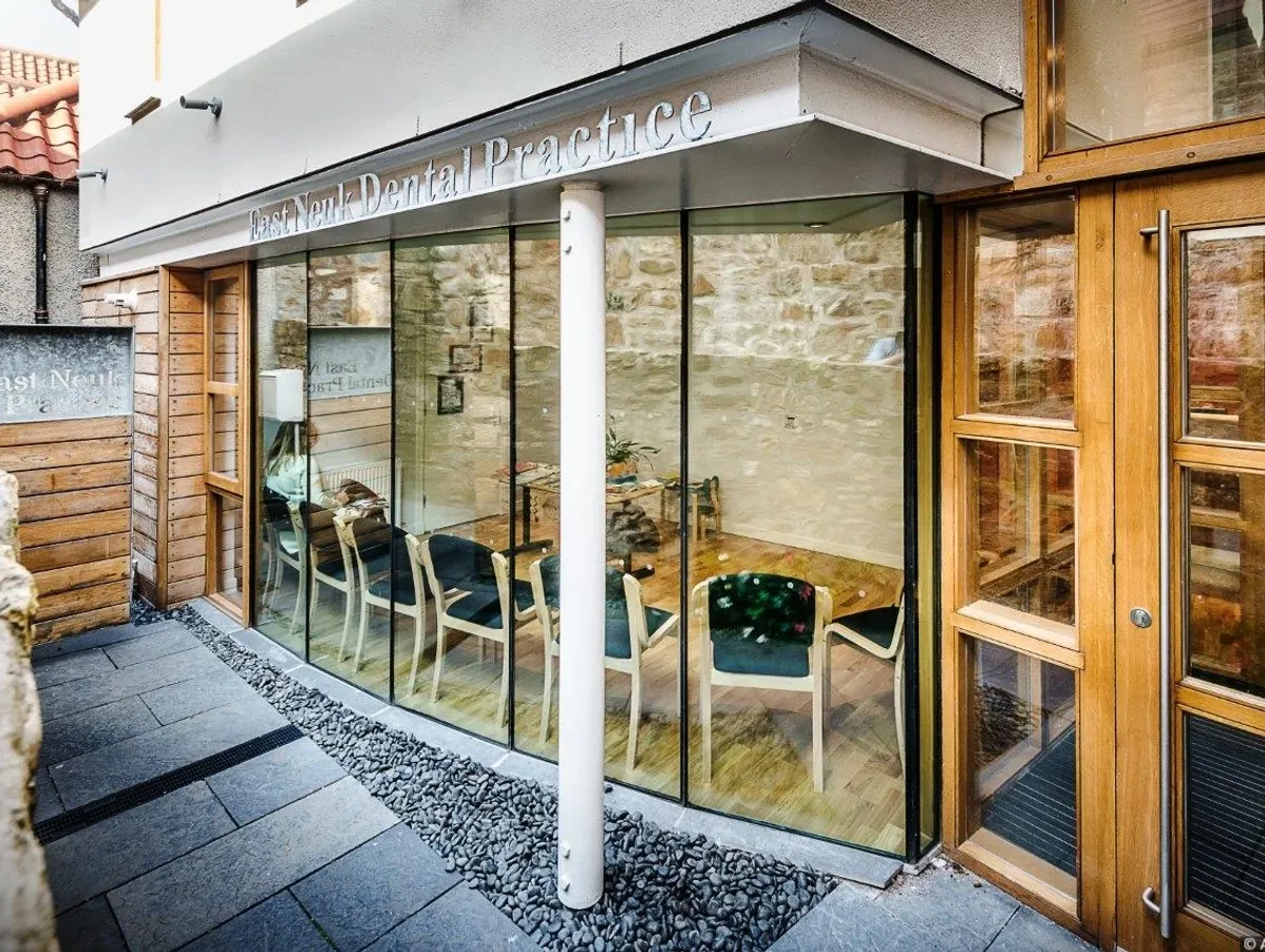 Exterior view of East Neuk Dental Practice with large glass windows showing a waiting area with wooden chairs inside.