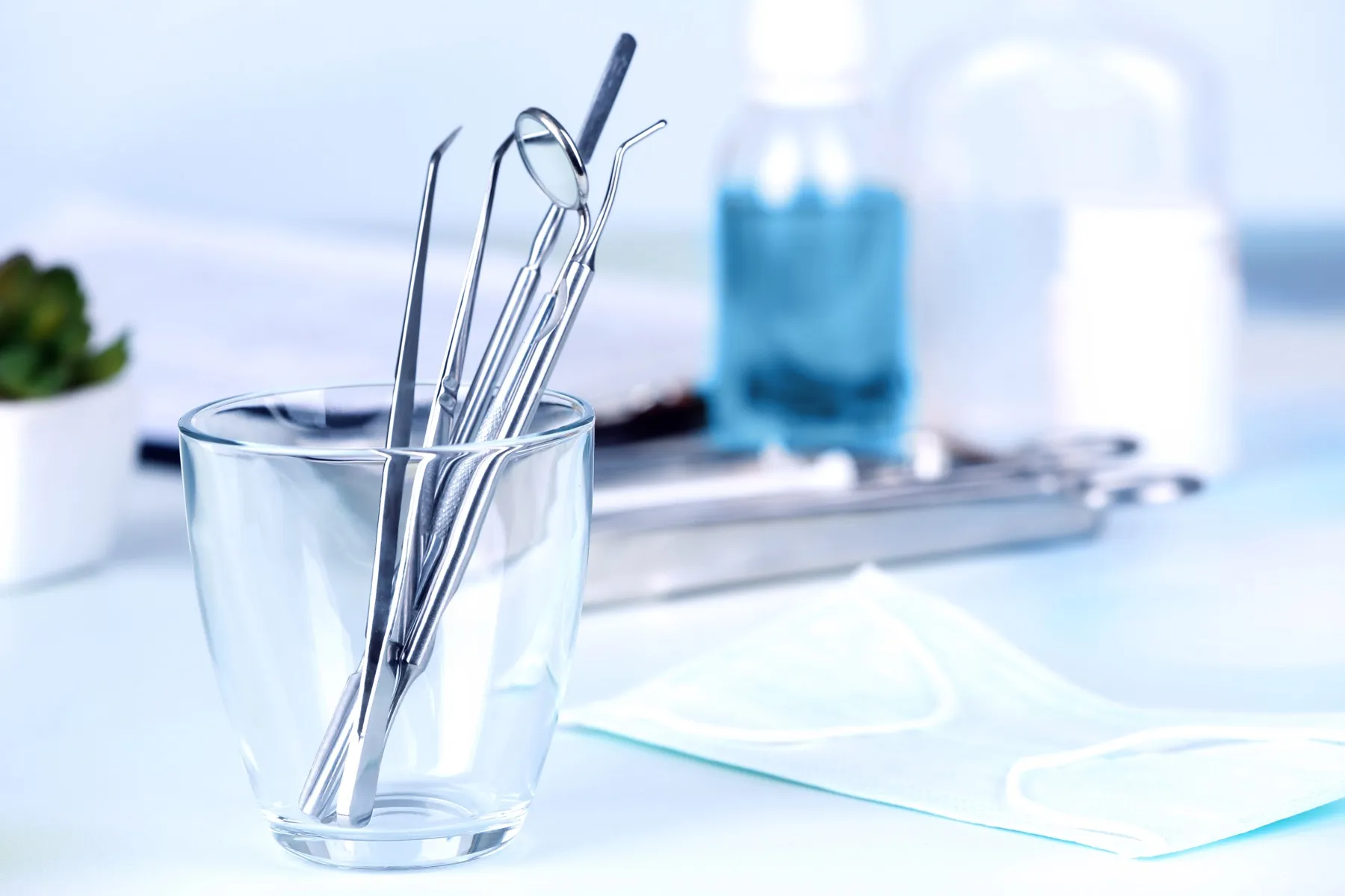 Dental tools including mirror, probe, and tweezers in a glass with a face mask and medical bottles on a table.