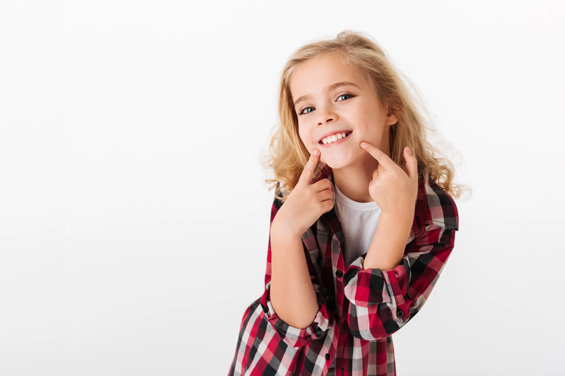 Smiling young girl in a red and black plaid shirt pointing to her teeth against white background.