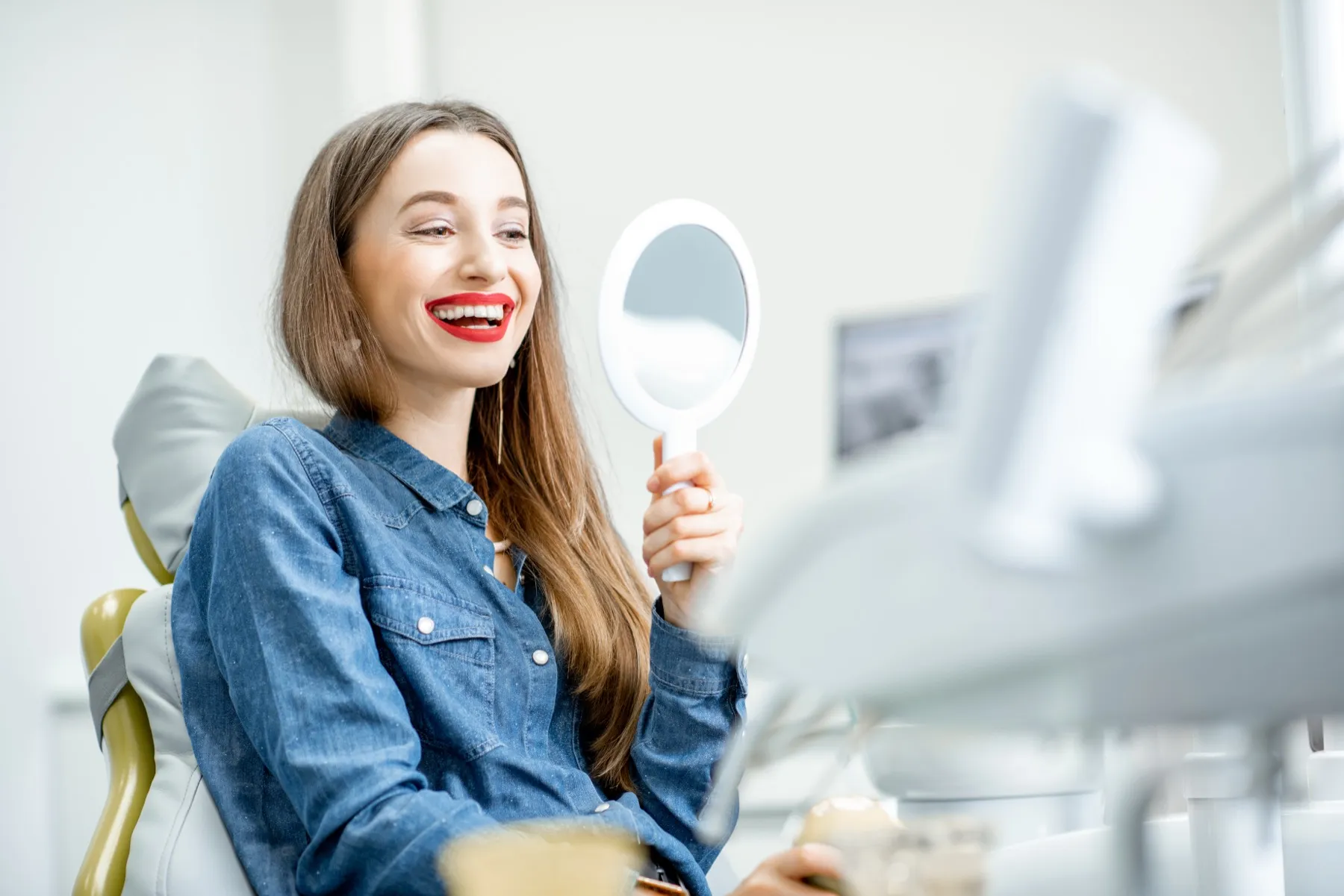 Smiling woman with red lipstick sitting in a dental chair, holding a round mirror and looking at her reflection.
