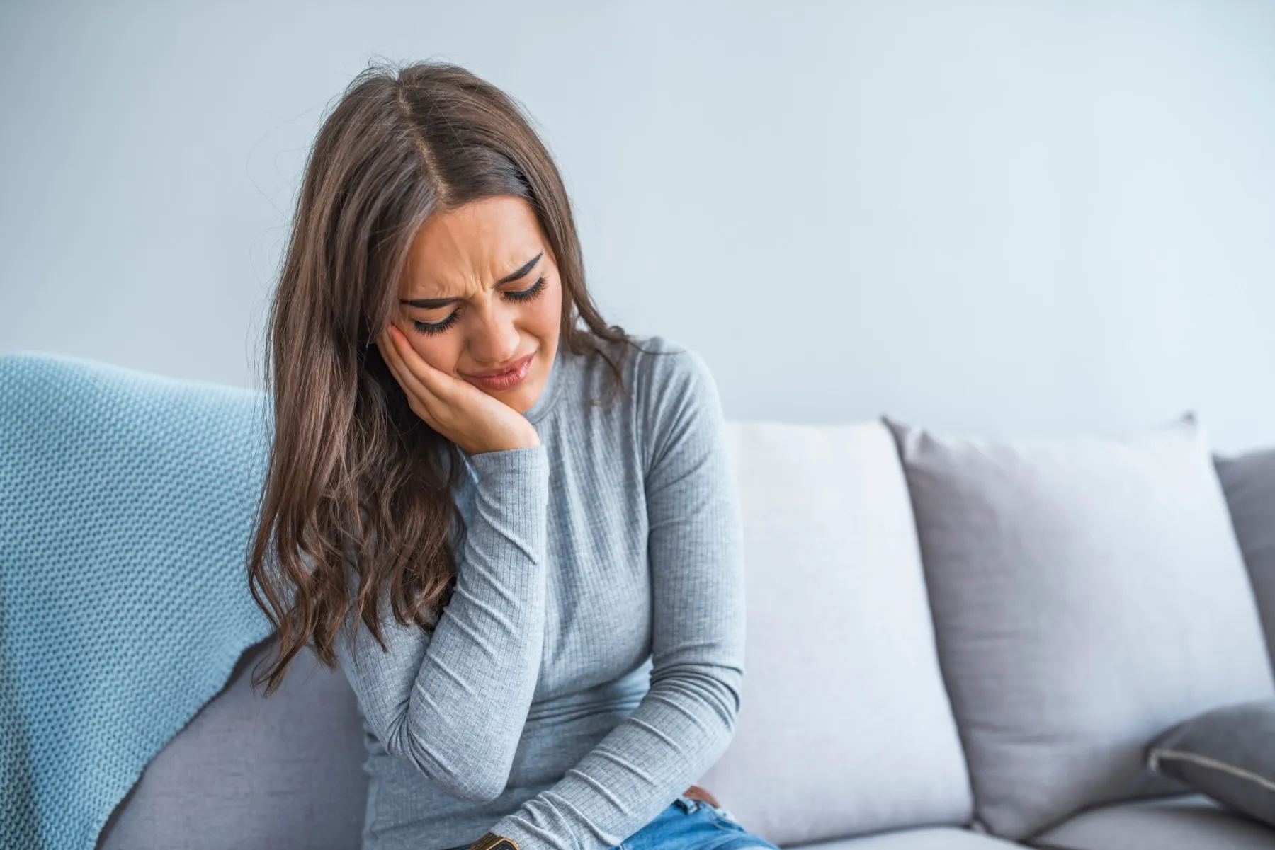 Young woman sitting on a couch holding her cheek with a pained expression, suggesting toothache or discomfort.