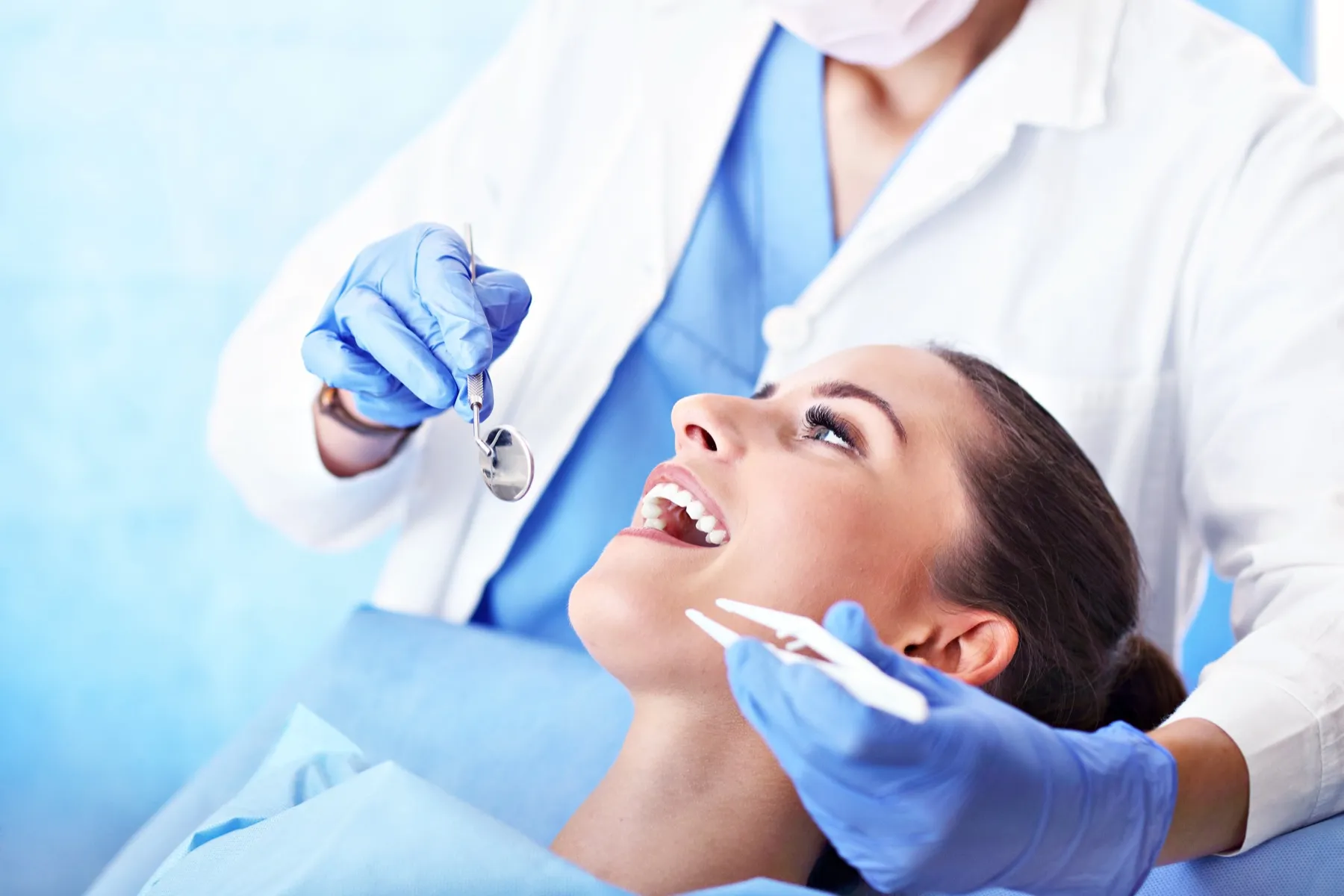 Dentist wearing blue gloves holding dental tools while examining a smiling female patient's teeth in a dental clinic.