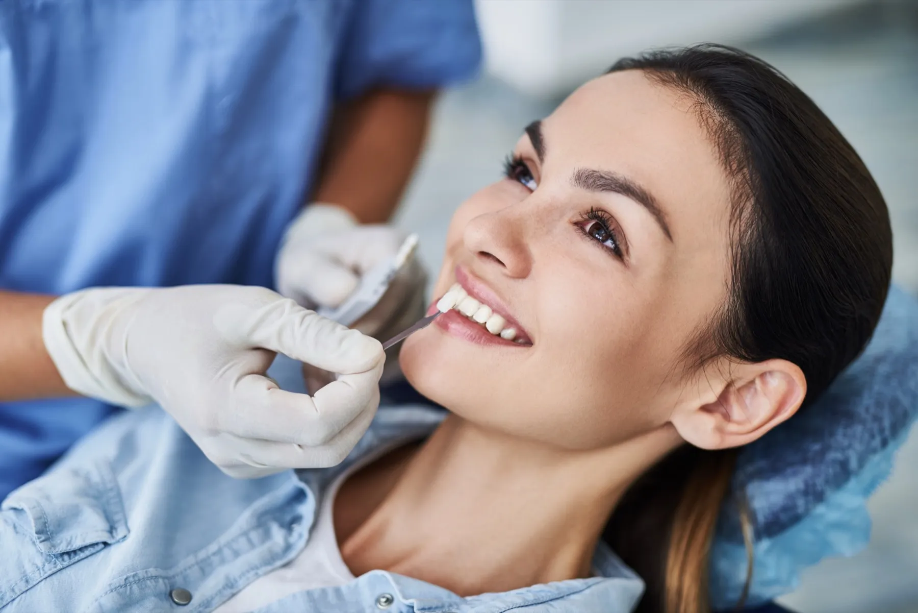 Dentist wearing white gloves comparing a shade guide to the teeth of a smiling woman in a dental chair.