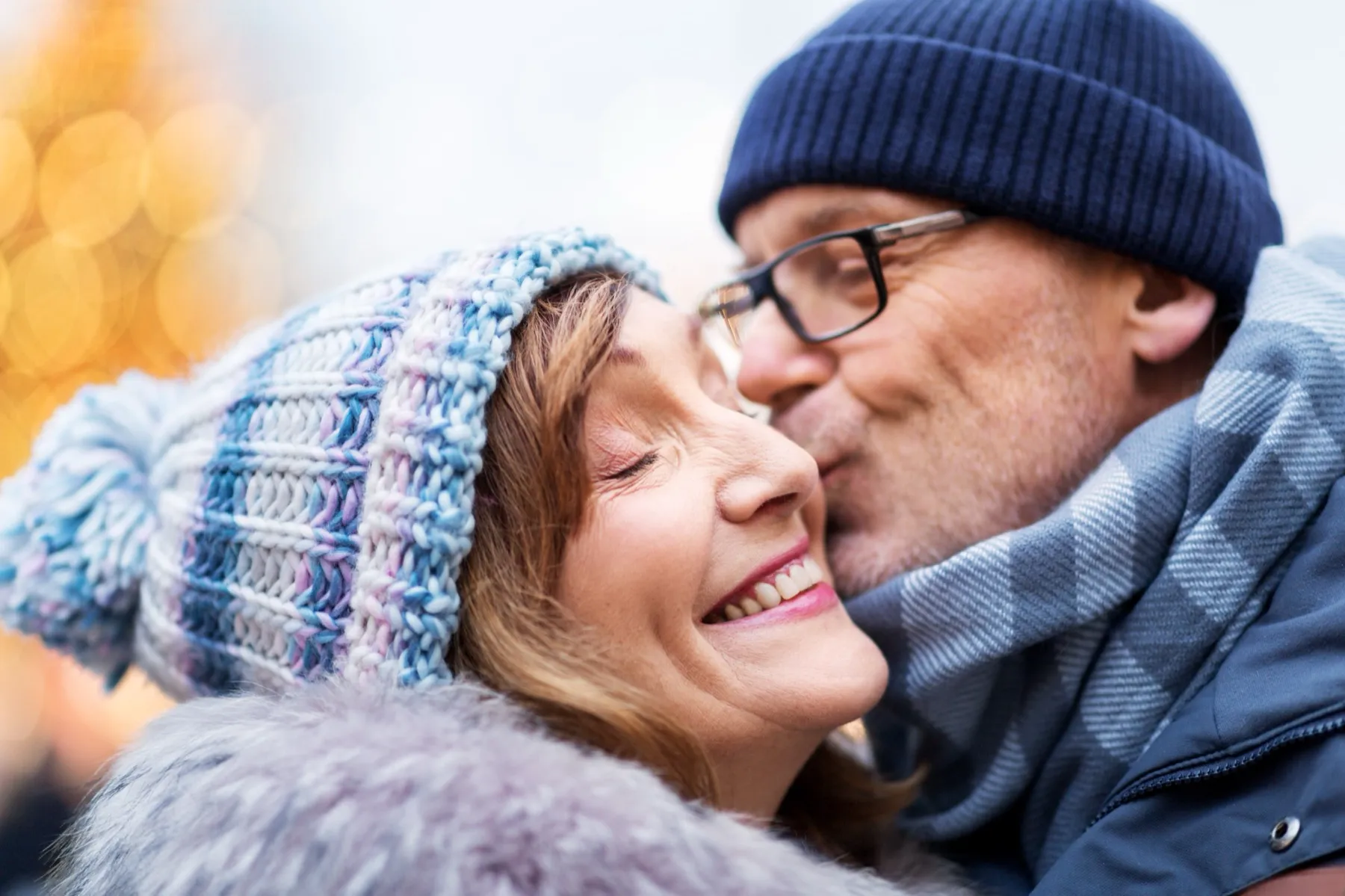 Older man wearing glasses and hat kissing smiling woman in a knitted winter hat on the cheek outdoors.