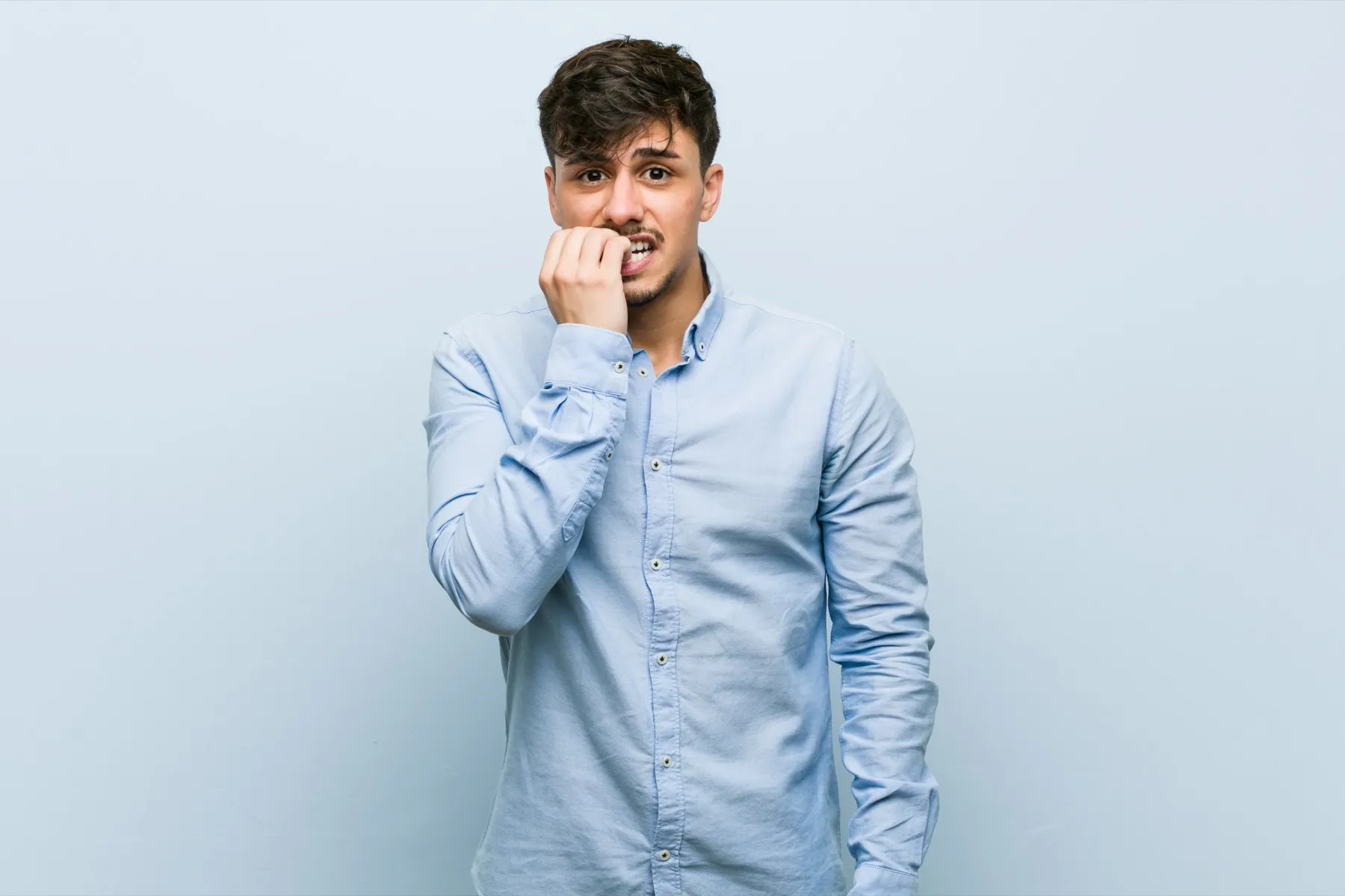 Nervous young man biting his nails wearing a light blue shirt against a pale background.