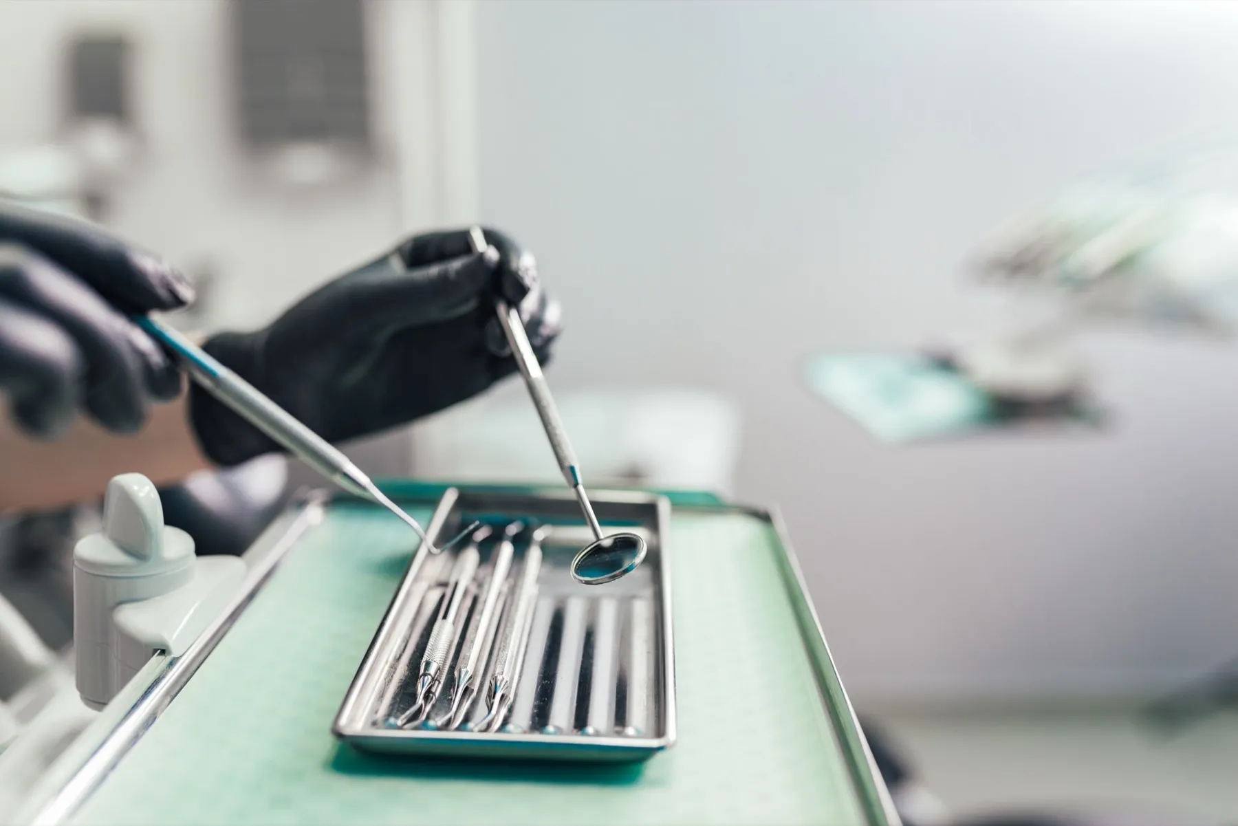 Close-up of a dentist's gloved hands holding dental mirror and probe over a tray of dental tools in a clinic.