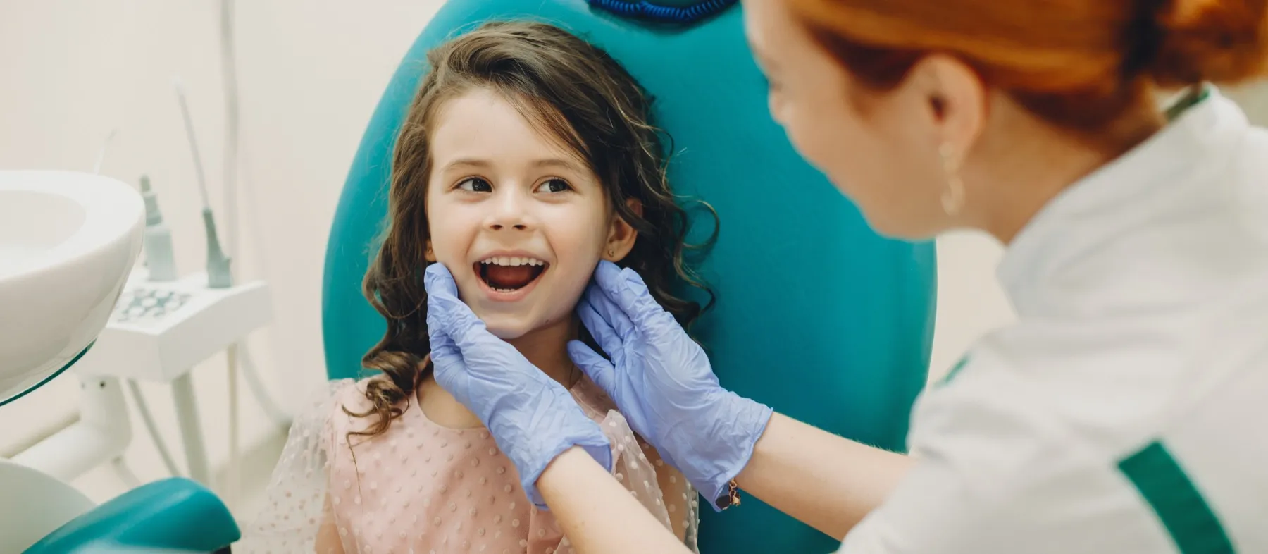 Dentist wearing blue gloves examines smiling young girl's teeth in dental chair.