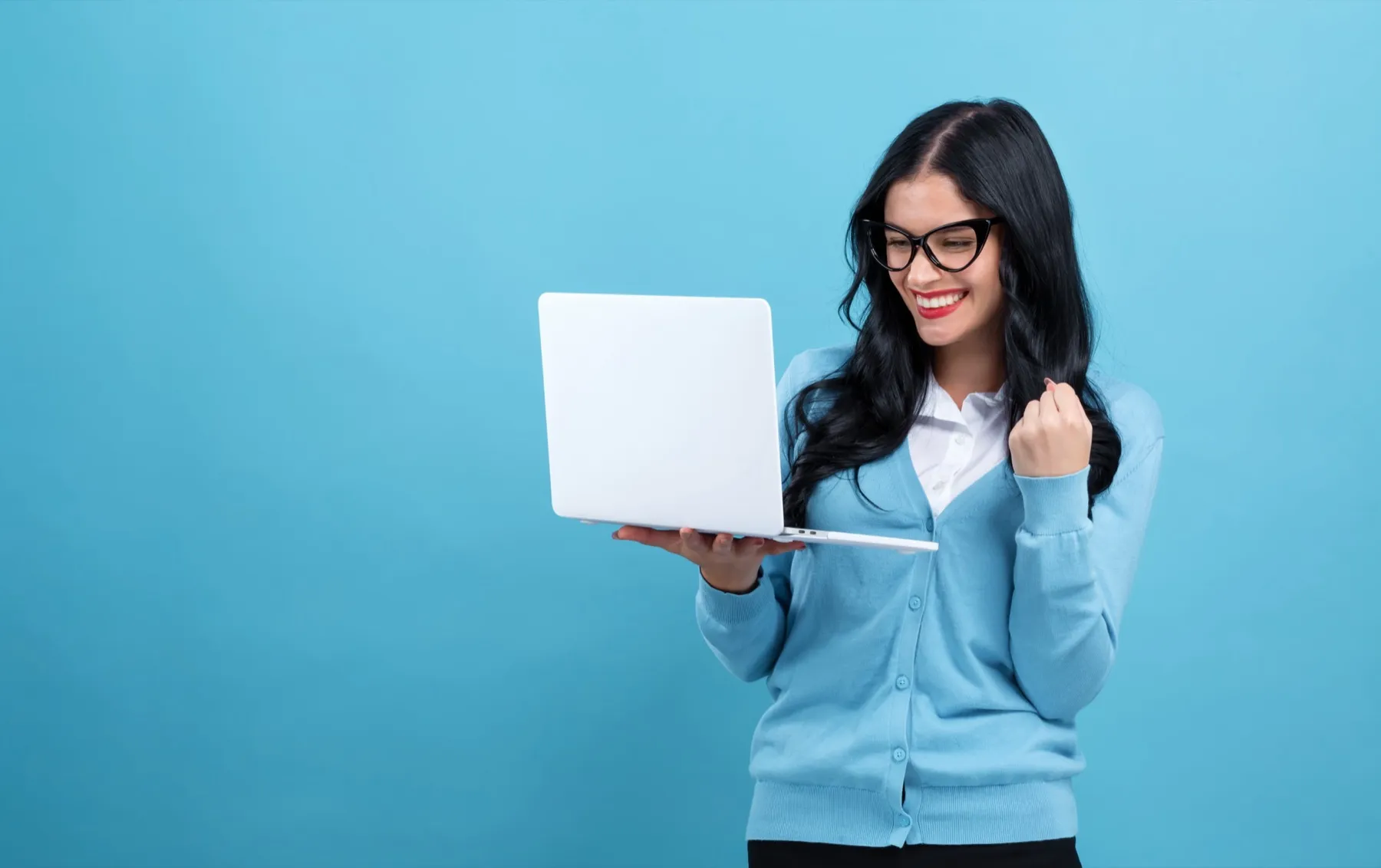 Smiling woman in glasses and blue cardigan holding a laptop and celebrating success against a blue background.